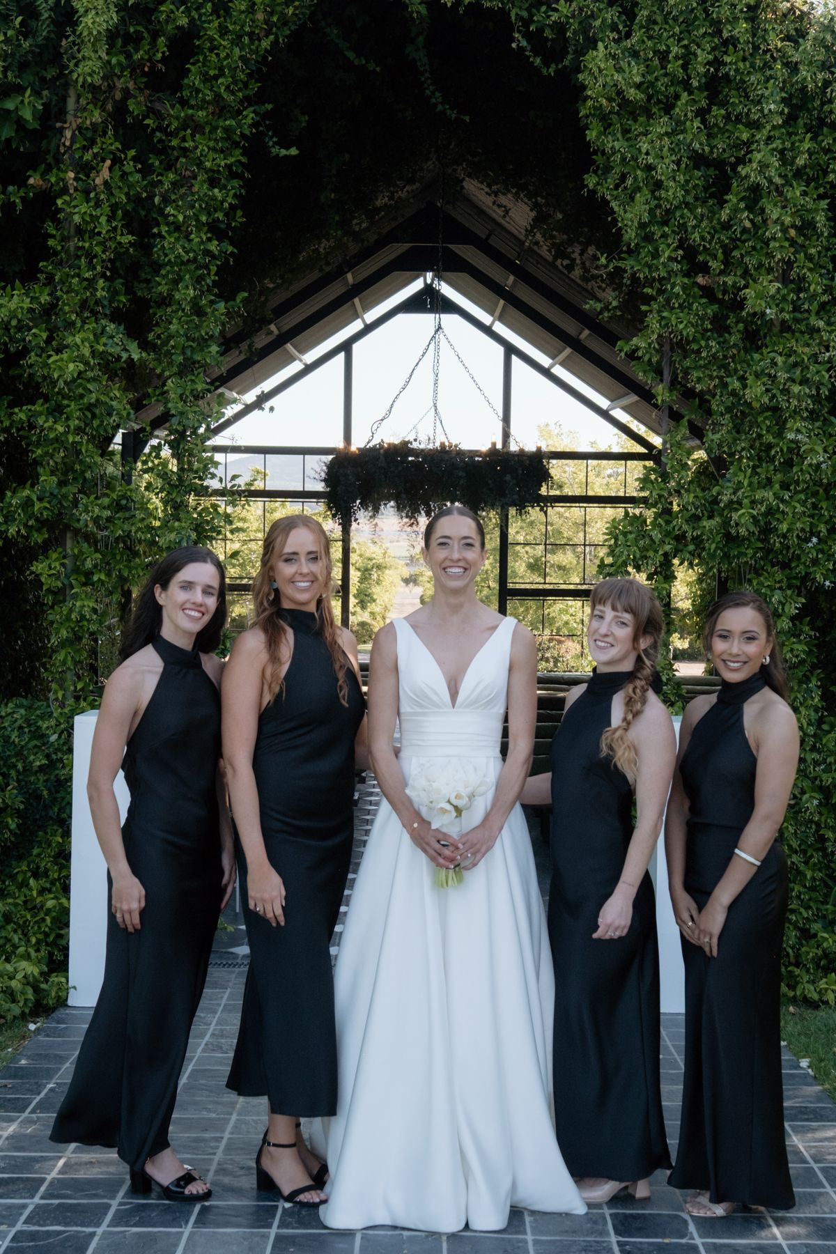 Bride in white V-neck gown poses with four bridesmaids in black halter dresses before glass chapel.