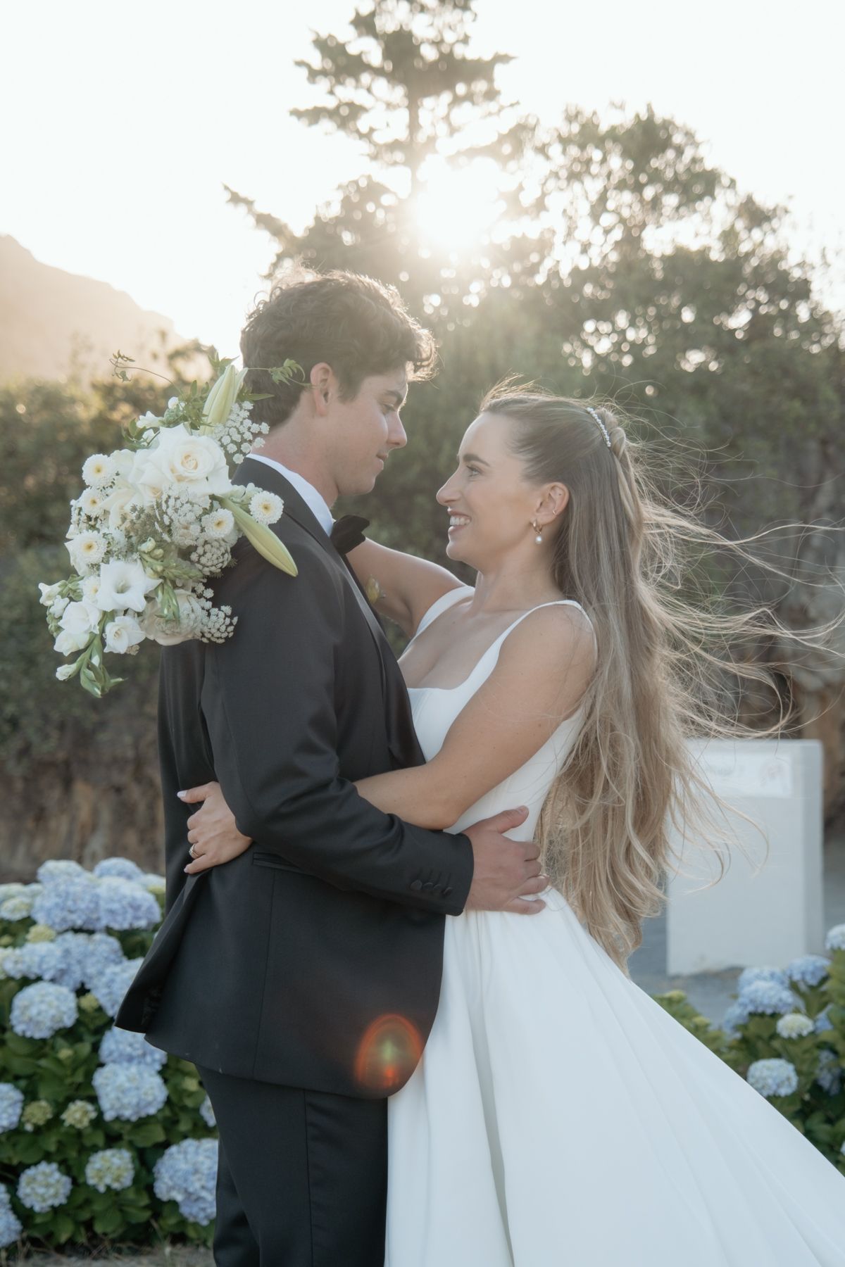 Bride and groom embracing at golden hour with white hydrangeas and bouquet.