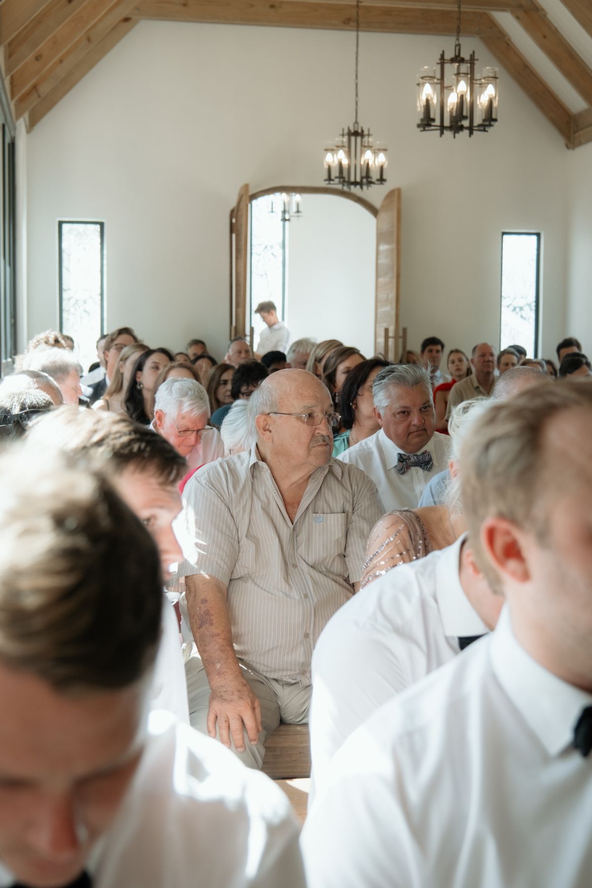 Guests seated in a bright chapel venue during a wedding ceremony, with chandeliers and arched doorway visible.