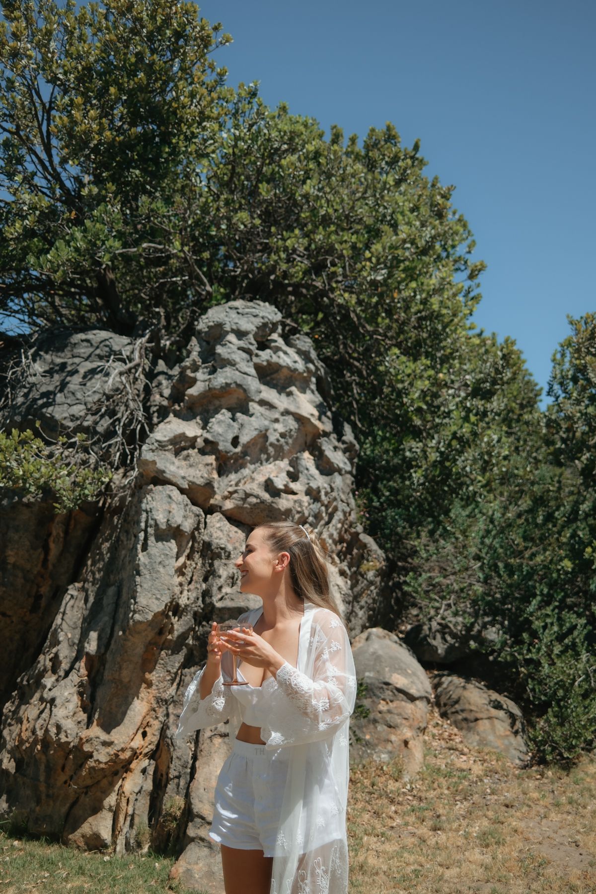 Bride in white lace robe standing near large rocks and green trees on a sunny day.