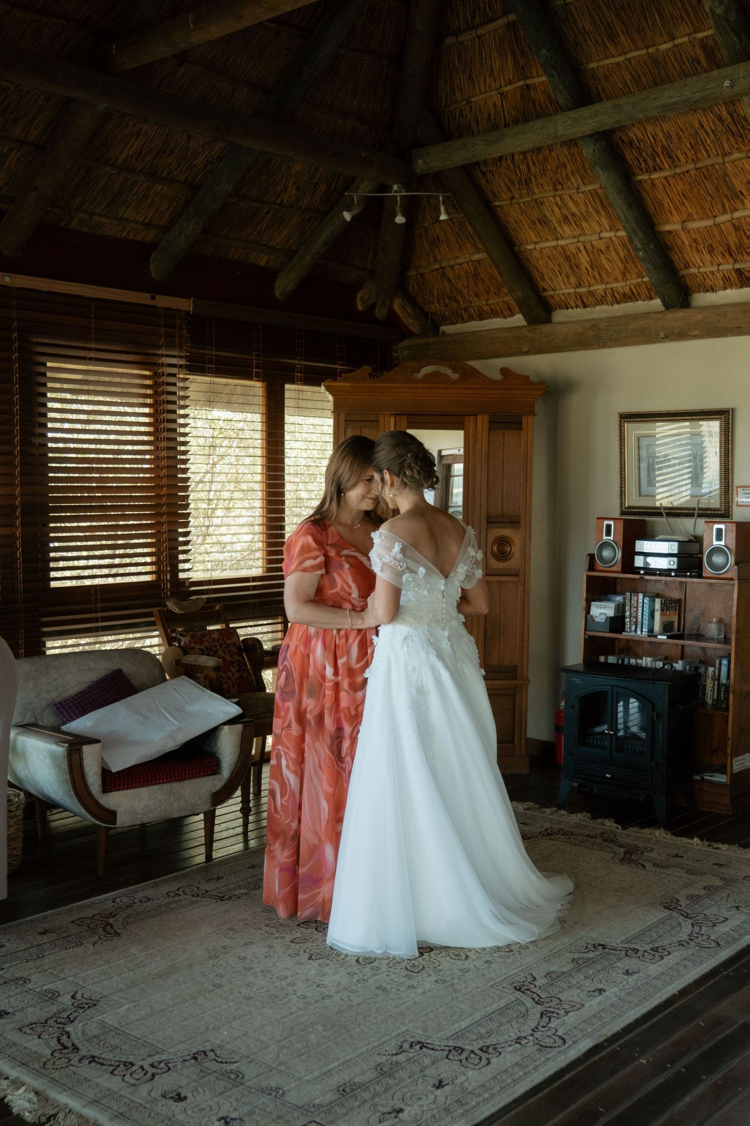 Bridesmaid in floral dress helping bride with her white lace gown in a rustic thatched interior.