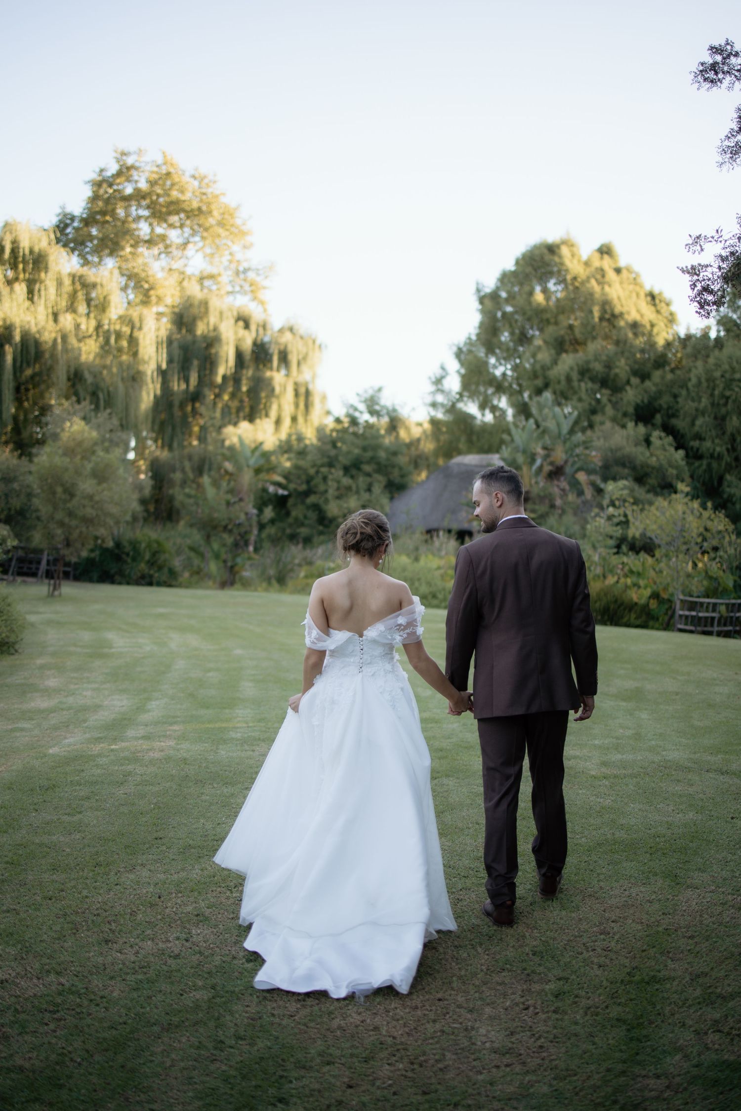 Bride and groom walking hand in hand across green lawn toward weeping willow trees at golden hour.