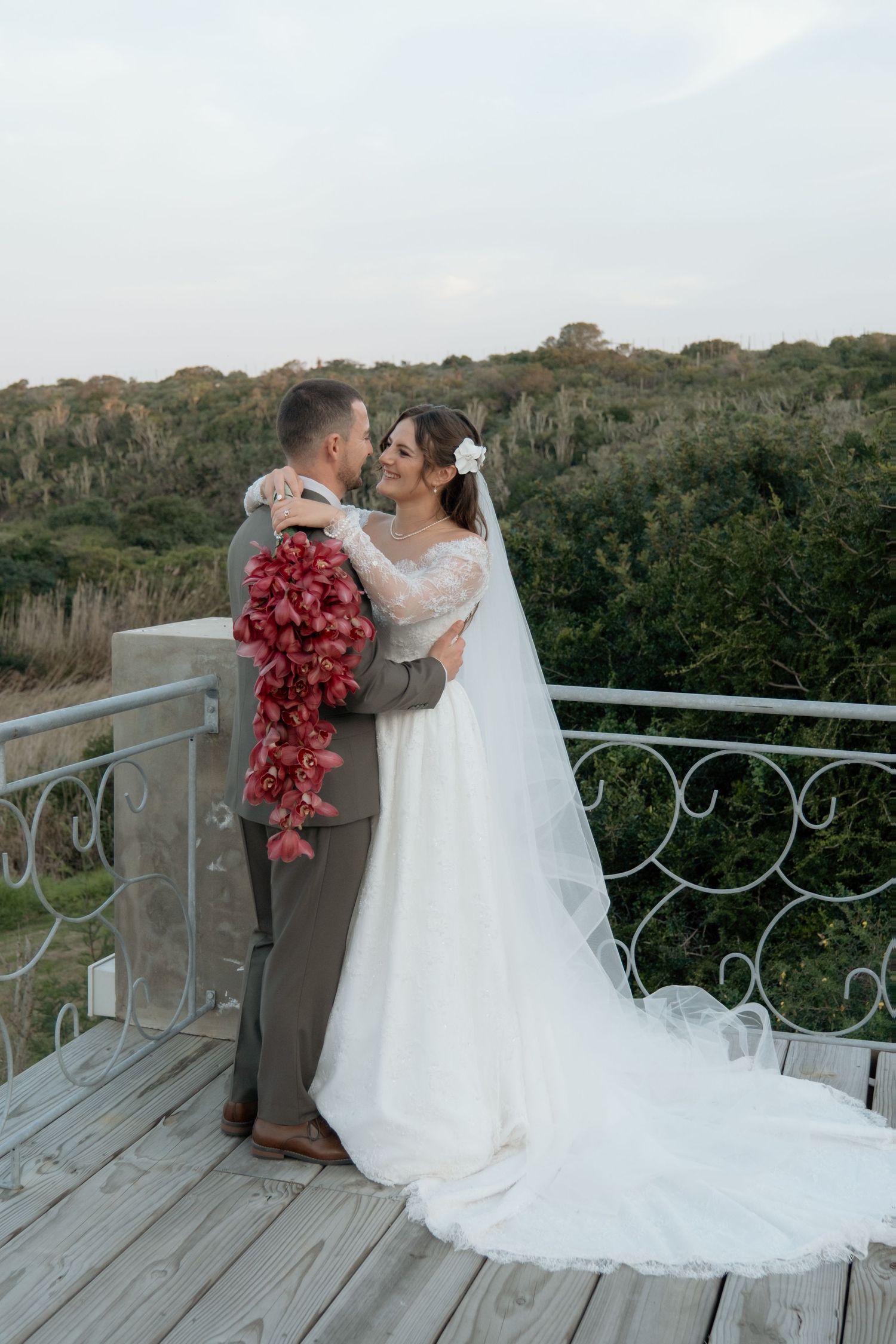 Bride and groom embracing on balcony with red cascading bouquet and scenic greenery backdrop.