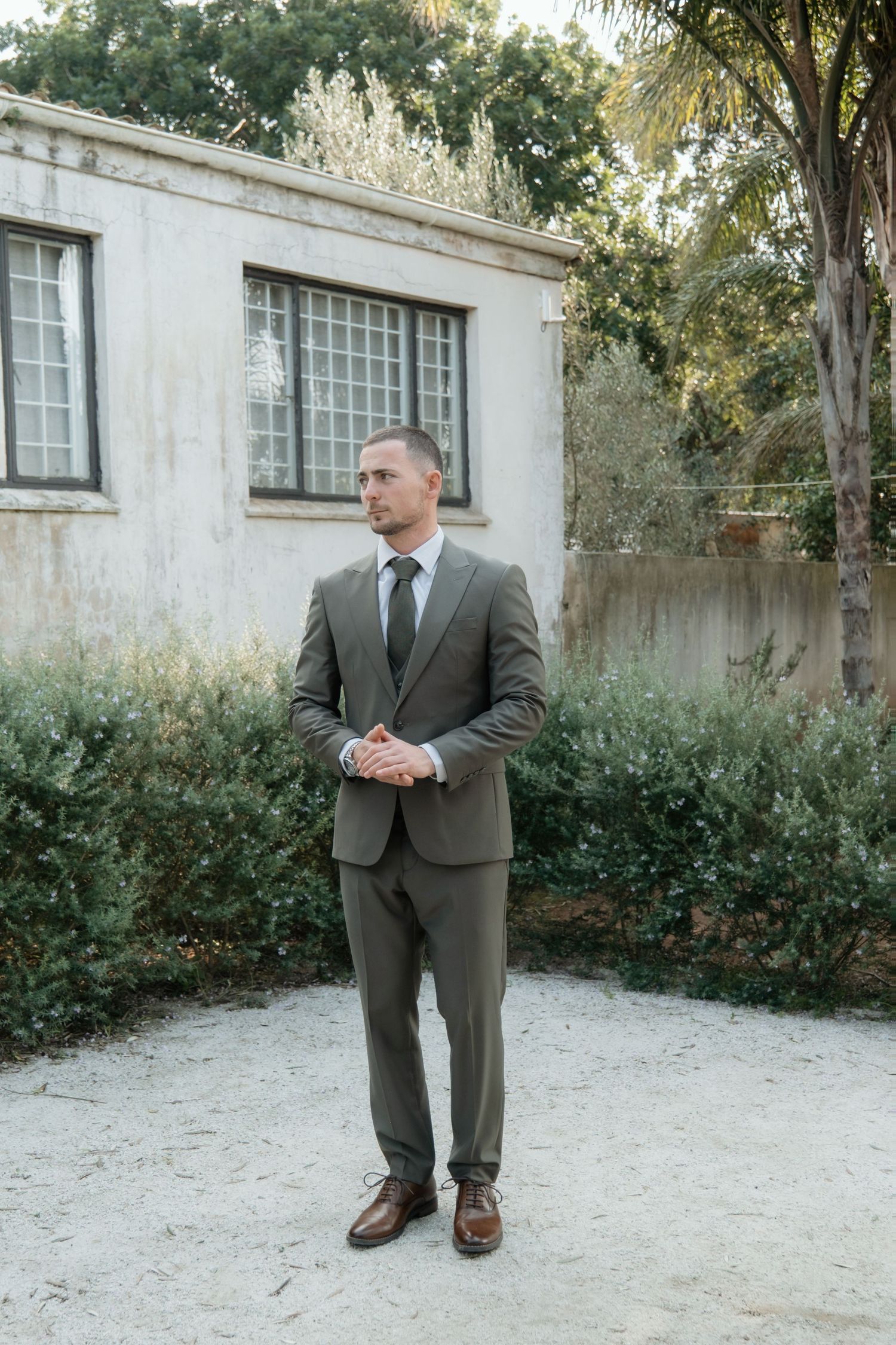 A man in a tailored olive suit and tie stands confidently outdoors near lush greenery and a stone building.