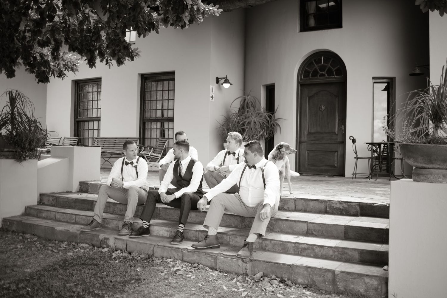 Black and white photo of groomsmen sitting on stone steps outside a classic white building before a wedding ceremony.