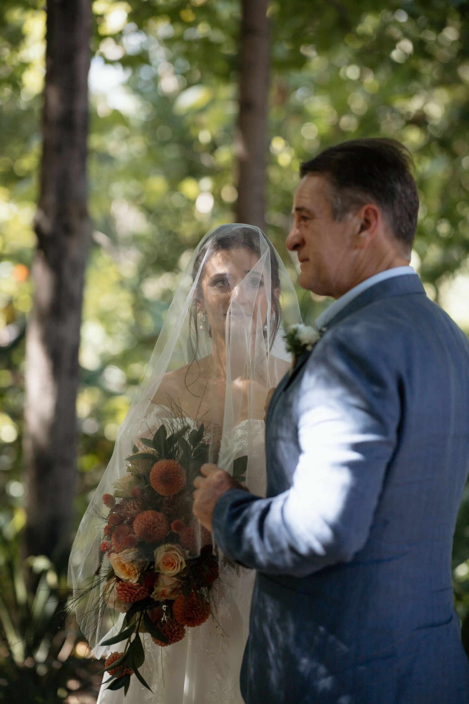 Bride wearing veil holding colorful bouquet standing beside father in blue suit in garden setting.