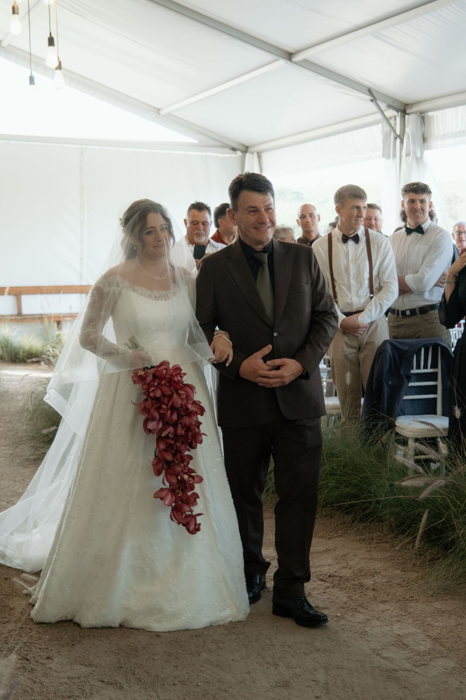 Bride in lace gown with red cascading bouquet walks down aisle with groom at outdoor tent wedding.