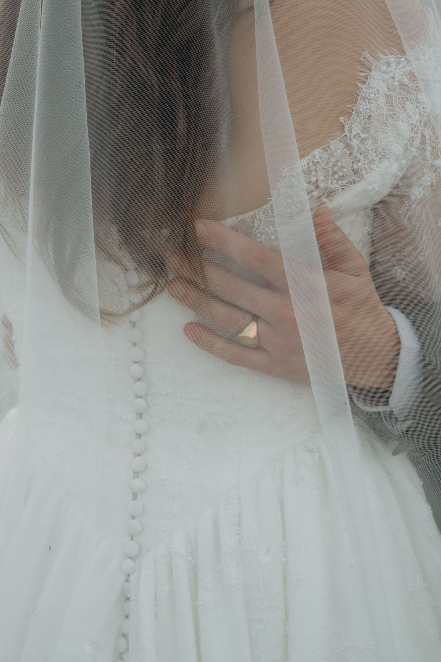 Groom placing hand on brides lace wedding dress back with ring visible under sheer veil.