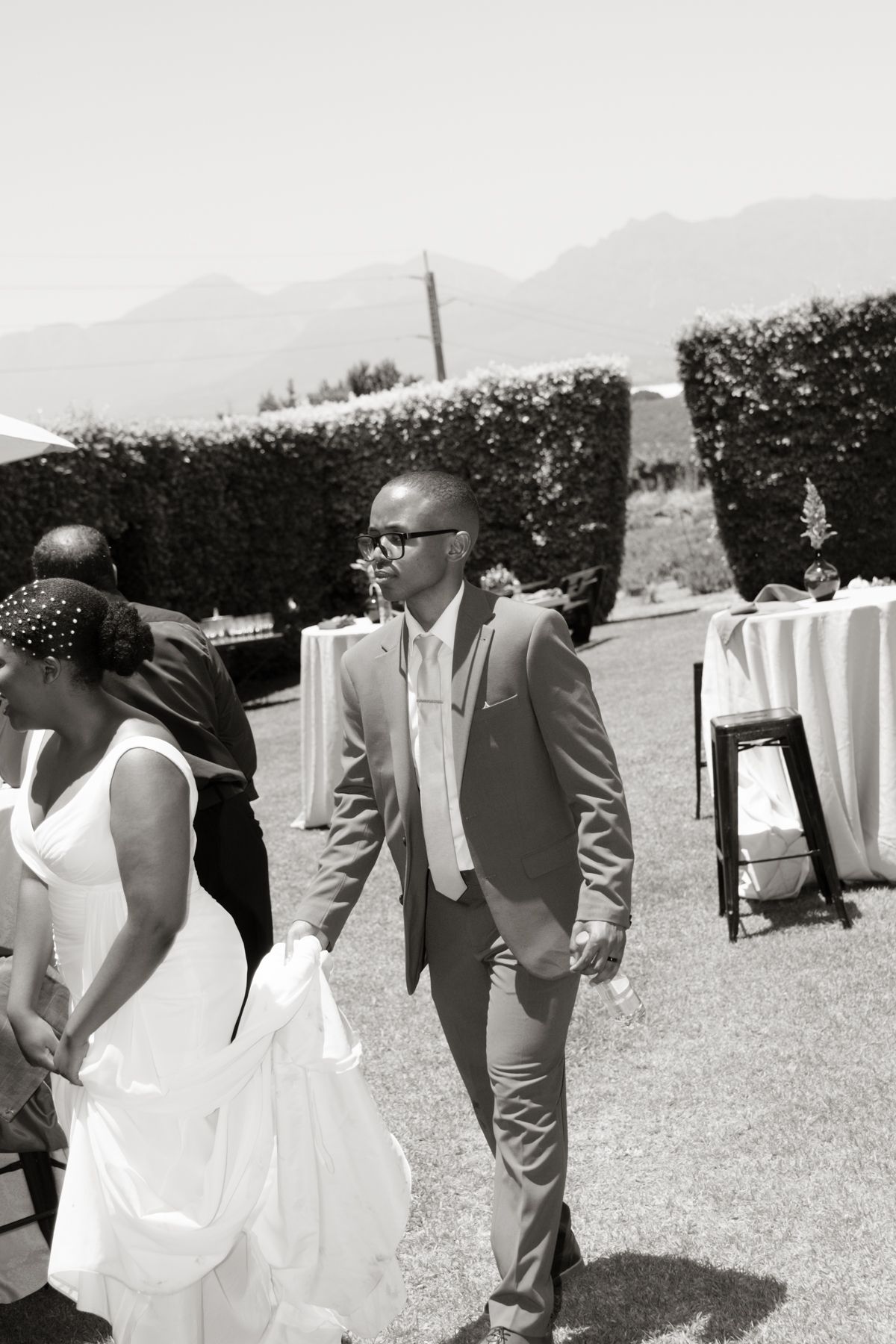 Black and white photo of a groom in a suit walking with a bride in a white dress at an outdoor wedding ceremony.