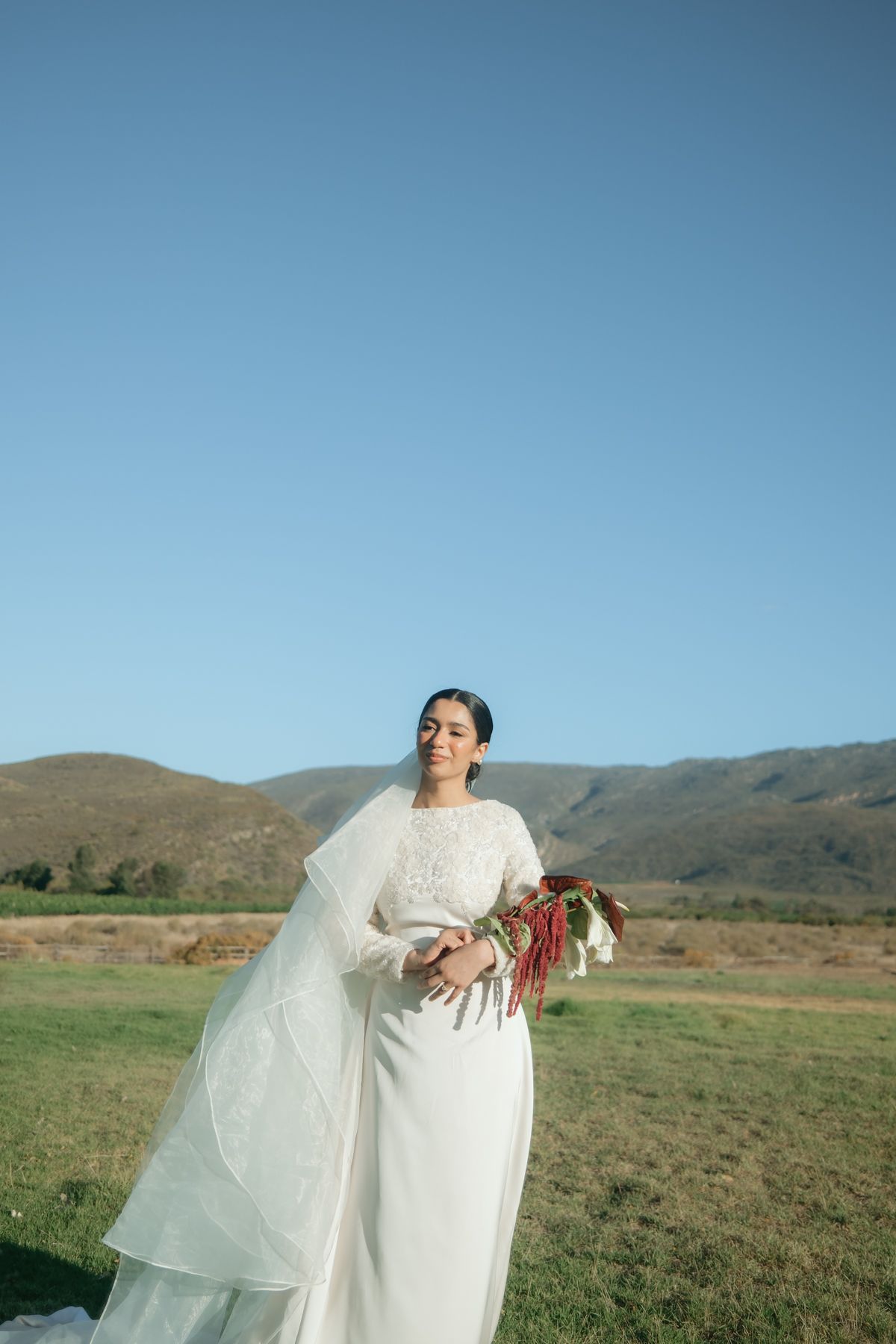 Smiling bride in white lace gown holds red bouquet in open field under blue sky.