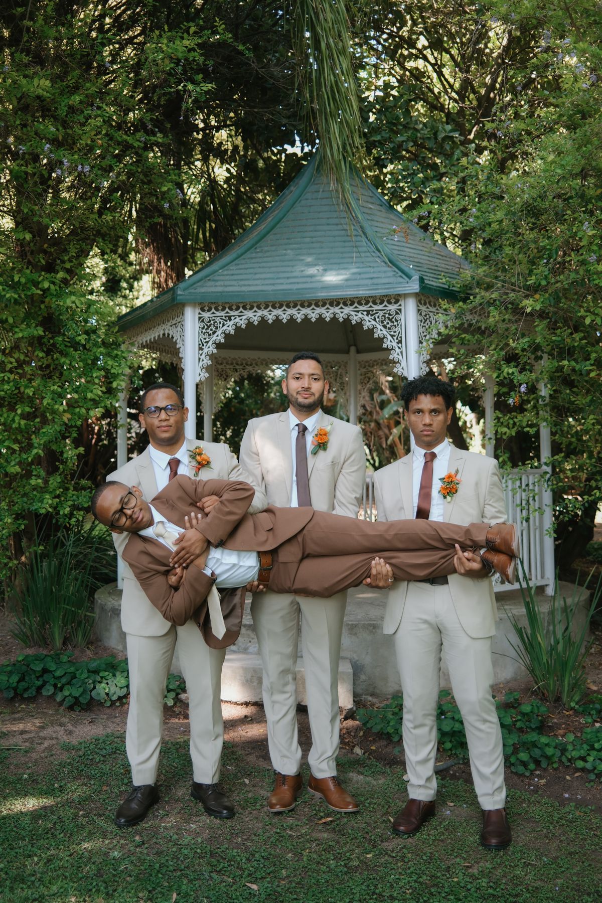 Three groomsmen playfully carry groom horizontally in front of white garden gazebo outdoors.