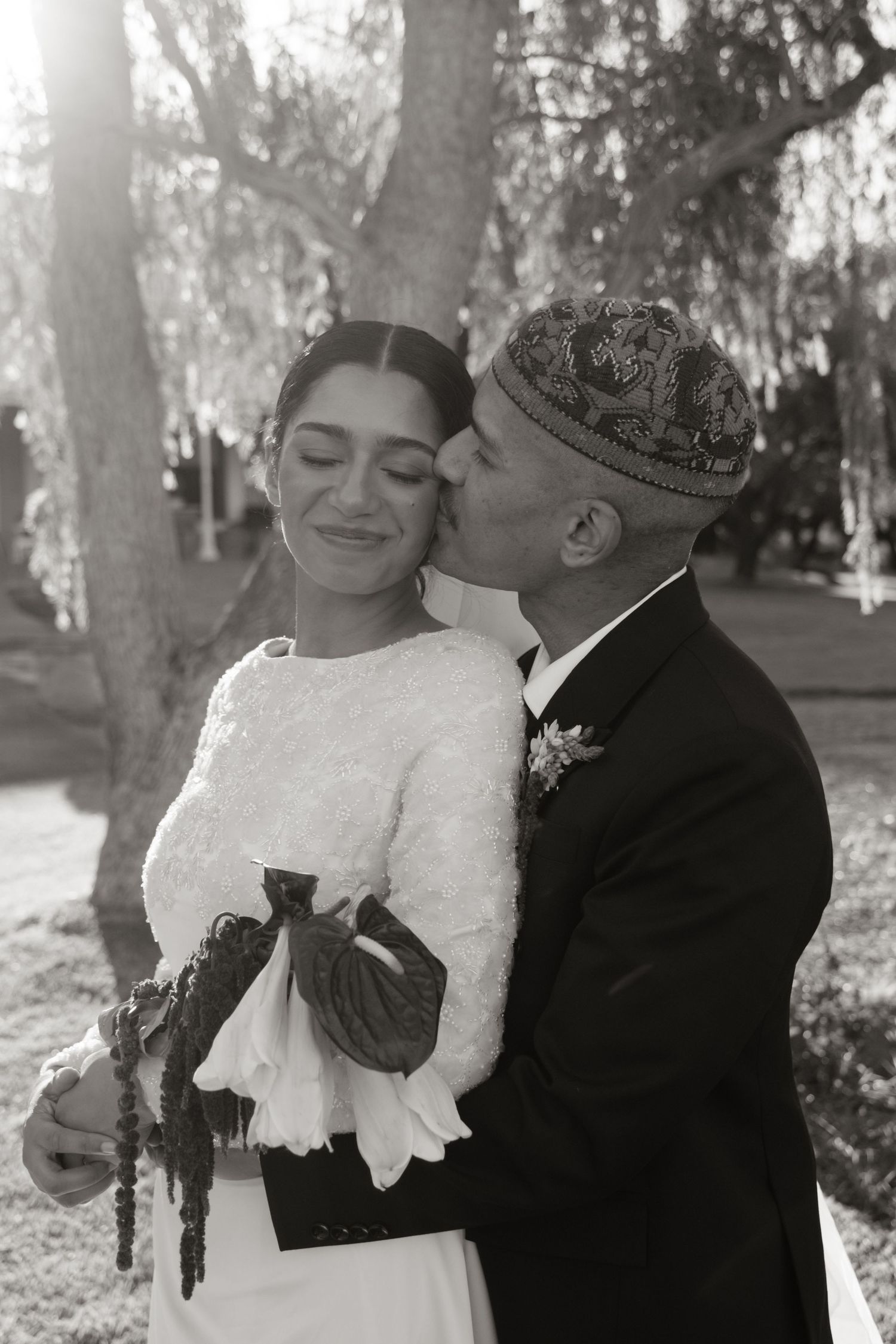 Black and white photo of groom kissing bride on cheek outdoors at wedding ceremony.