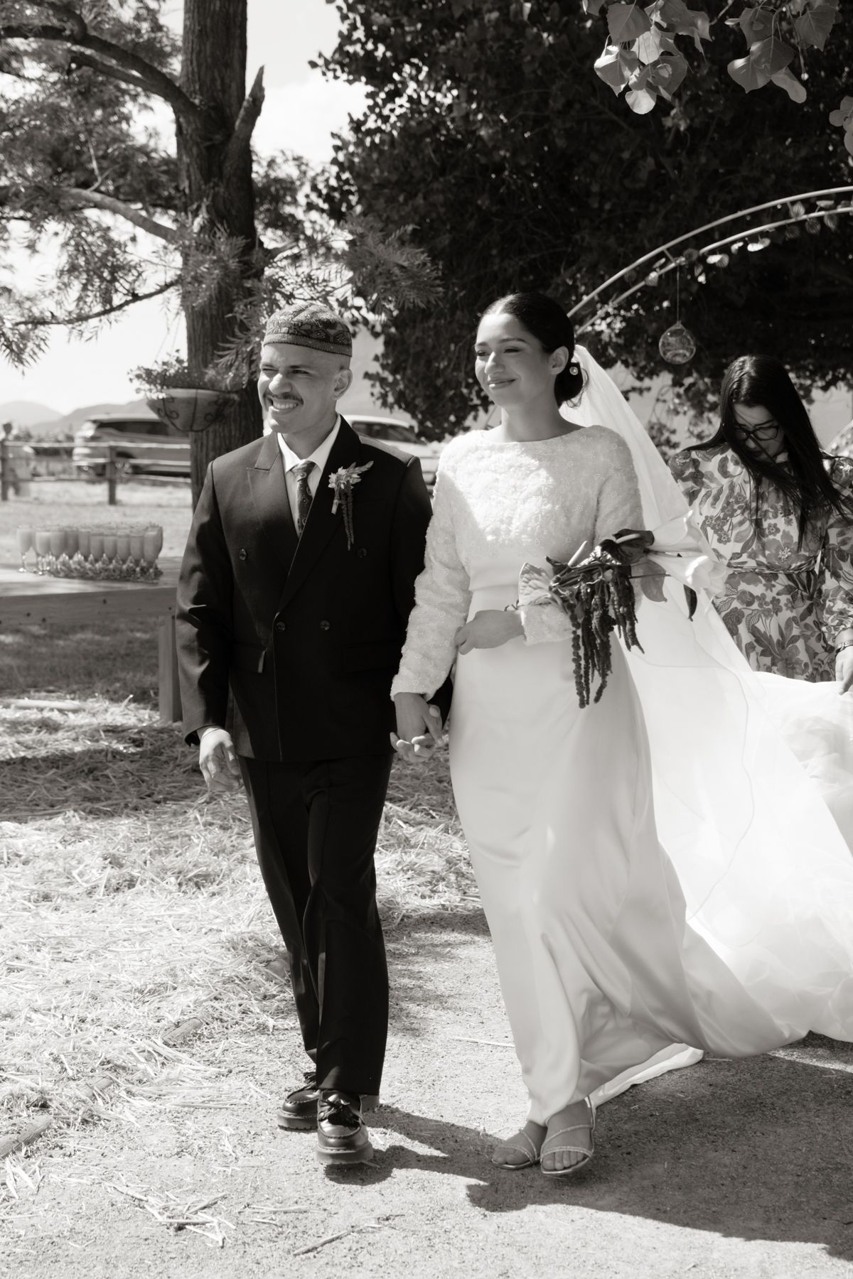 Bride and groom walking hand in hand in black and white wedding photo beneath shady trees.