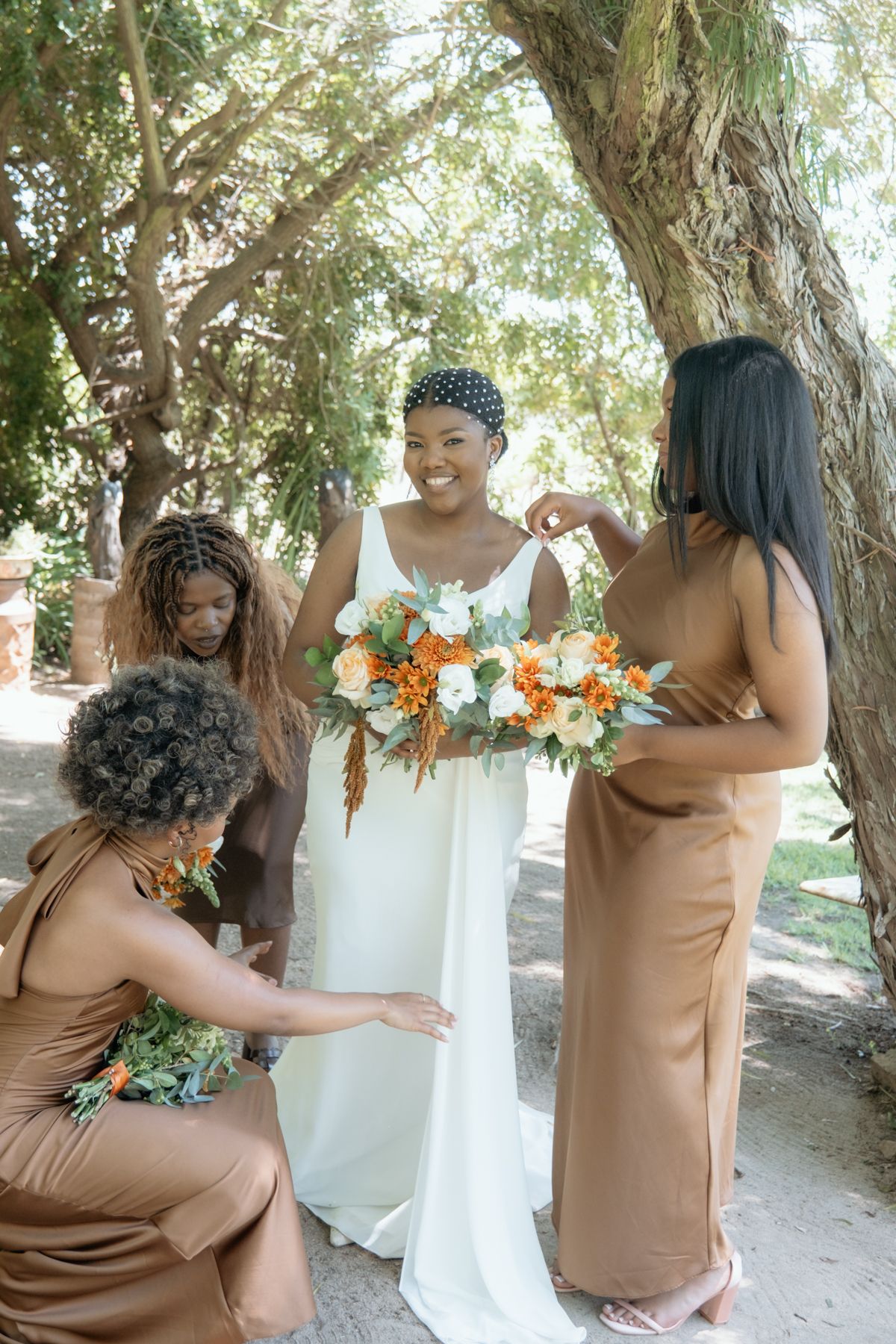 Bride in white gown smiles as bridesmaids in brown satin dresses help her with orange and white bouquets outdoors.