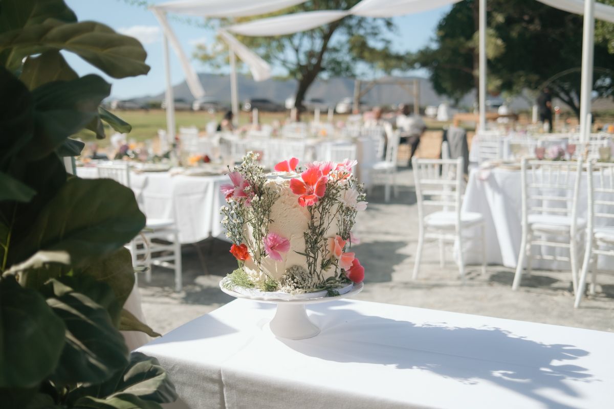 Elegant single-tier wedding cake with colorful floral decorations on white stand at outdoor reception.