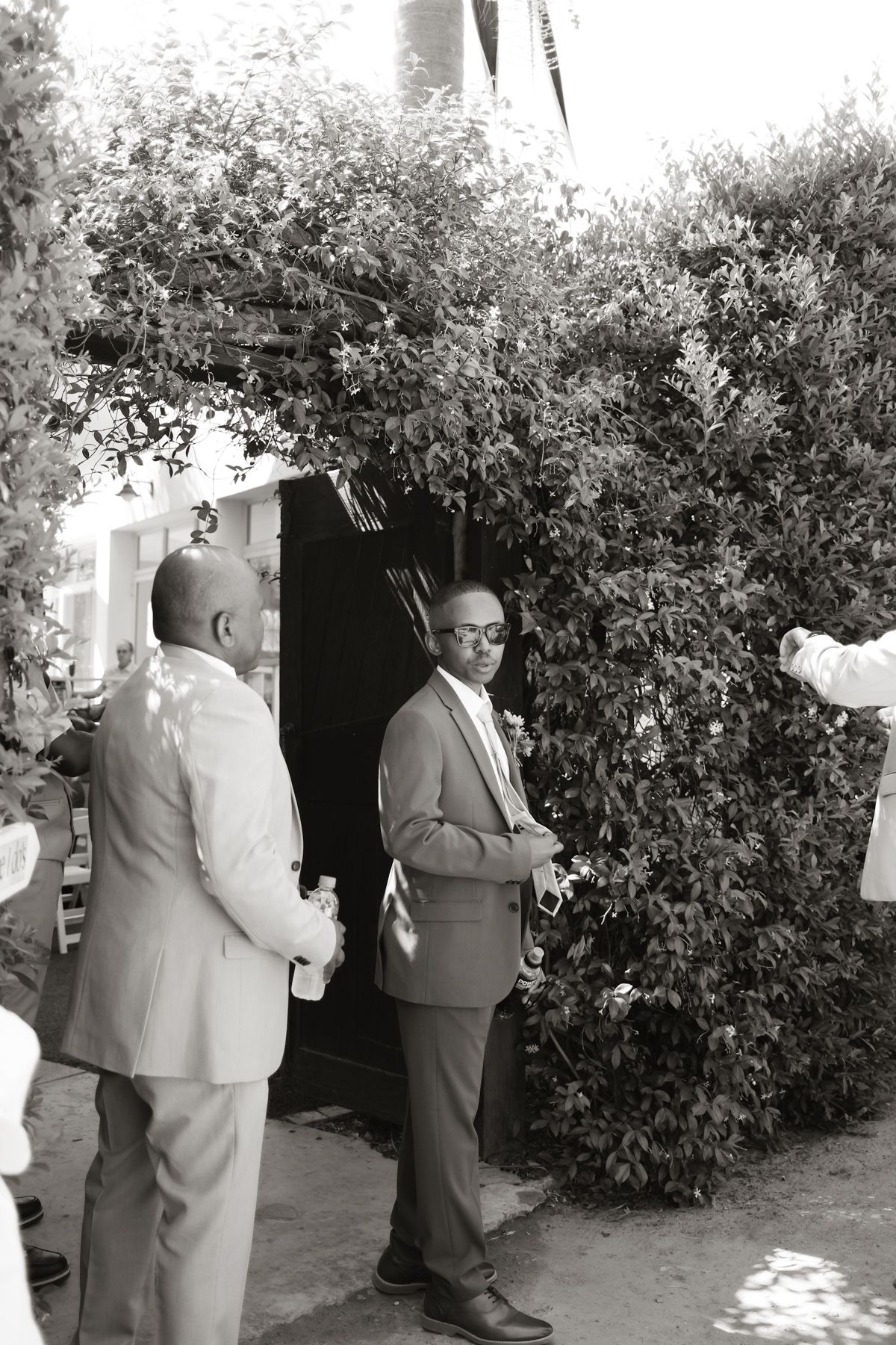 Two men in suits stand near a lush green hedge at an outdoor wedding venue.