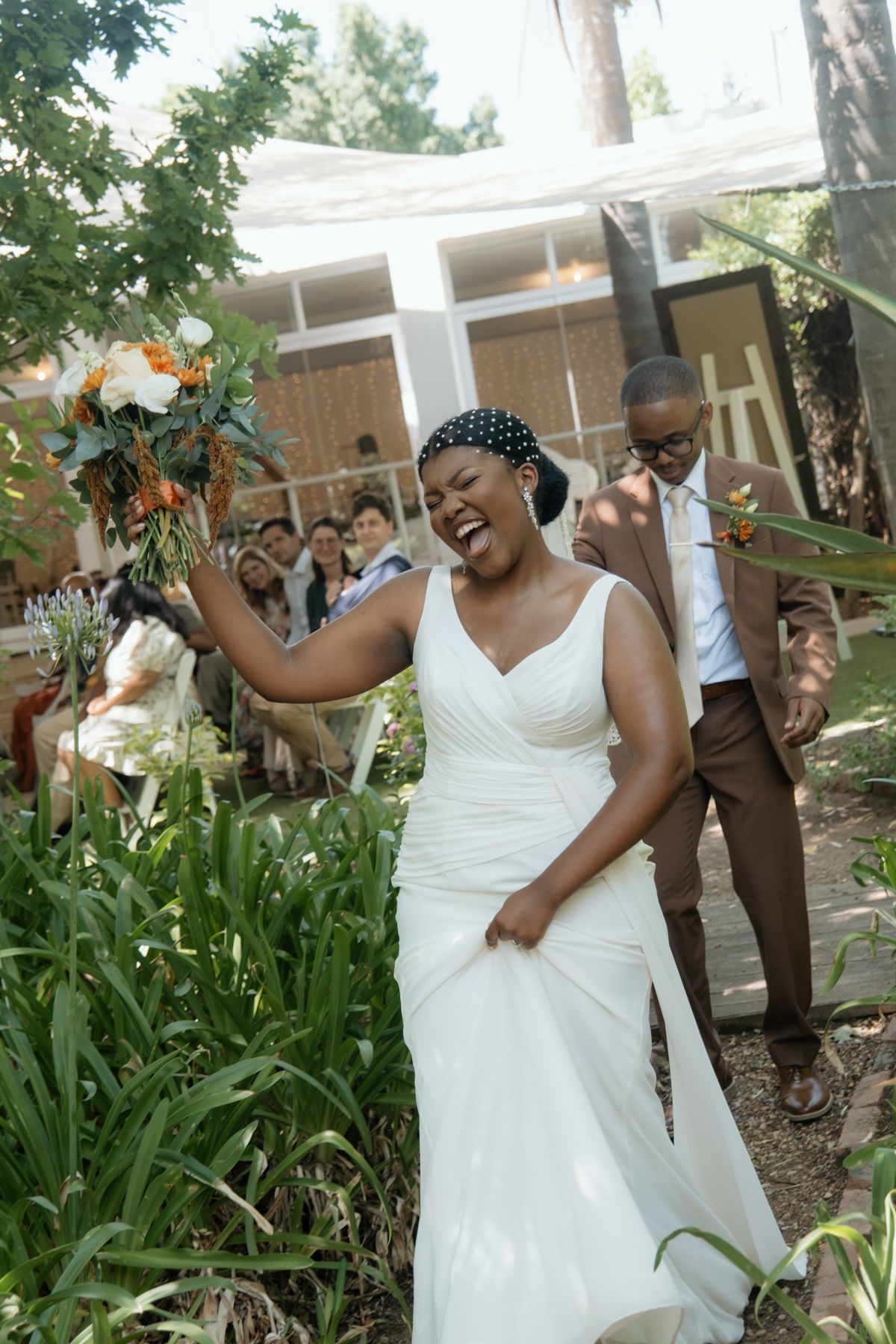 Elated Black bride laughs while raising bouquet of orange and white flowers after outdoor wedding ceremony.