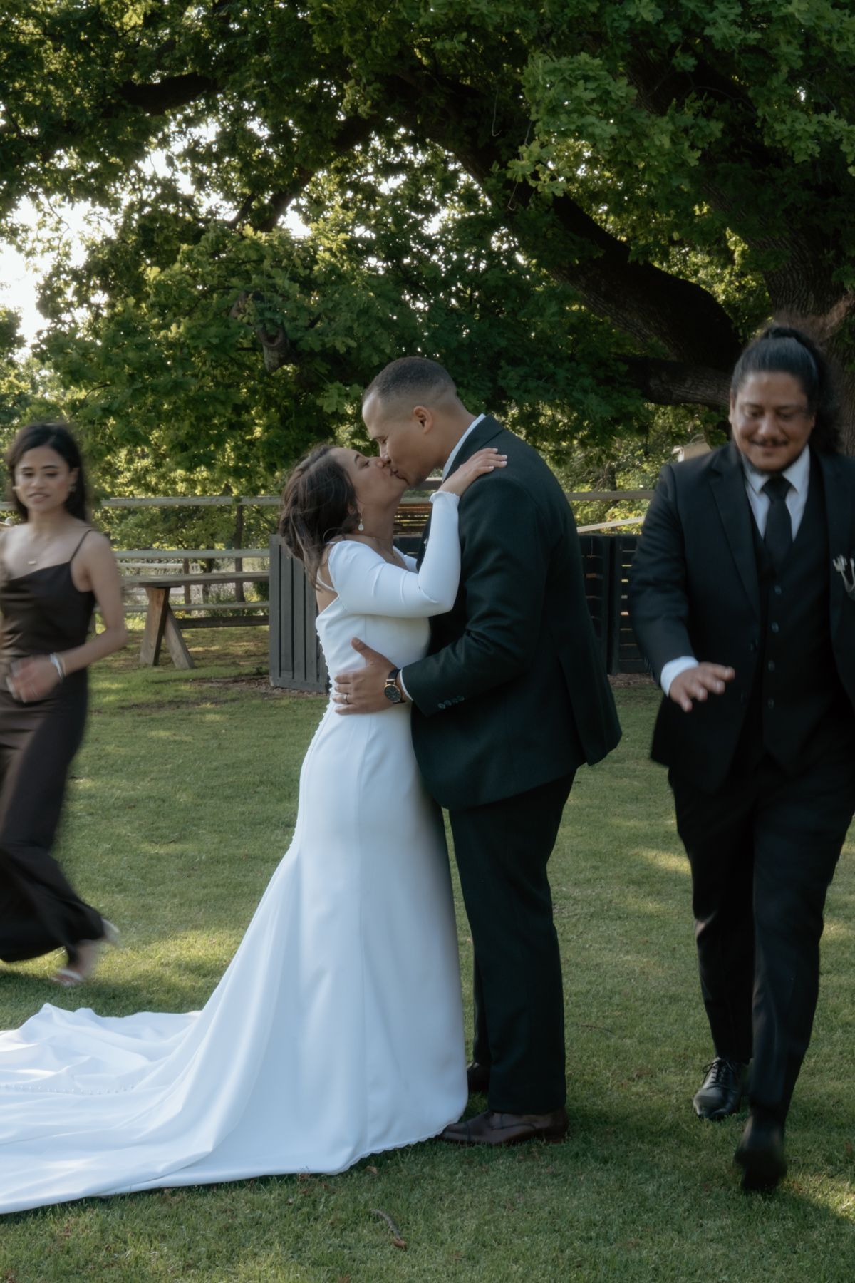 Bride and groom share a kiss outdoors on grass while wedding guests celebrate around them beneath a large tree.