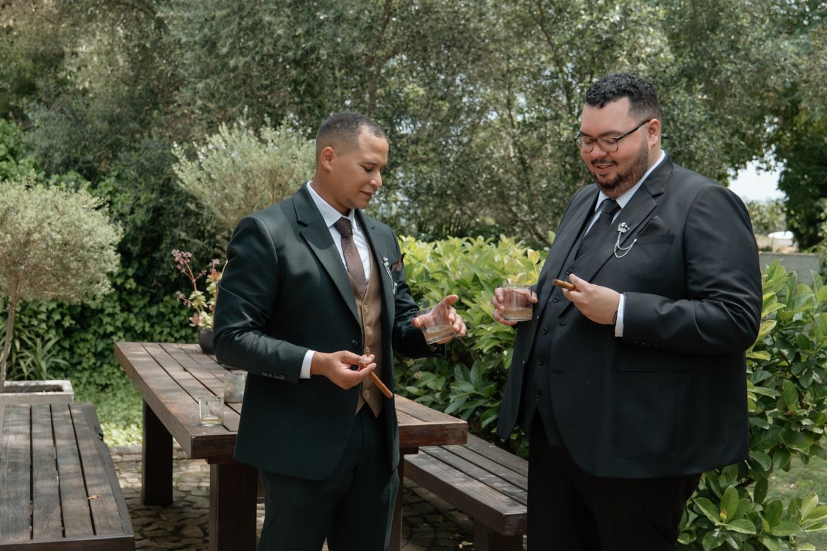 Two groomsmen in black suits sharing a laugh while holding drinks in a garden setting.