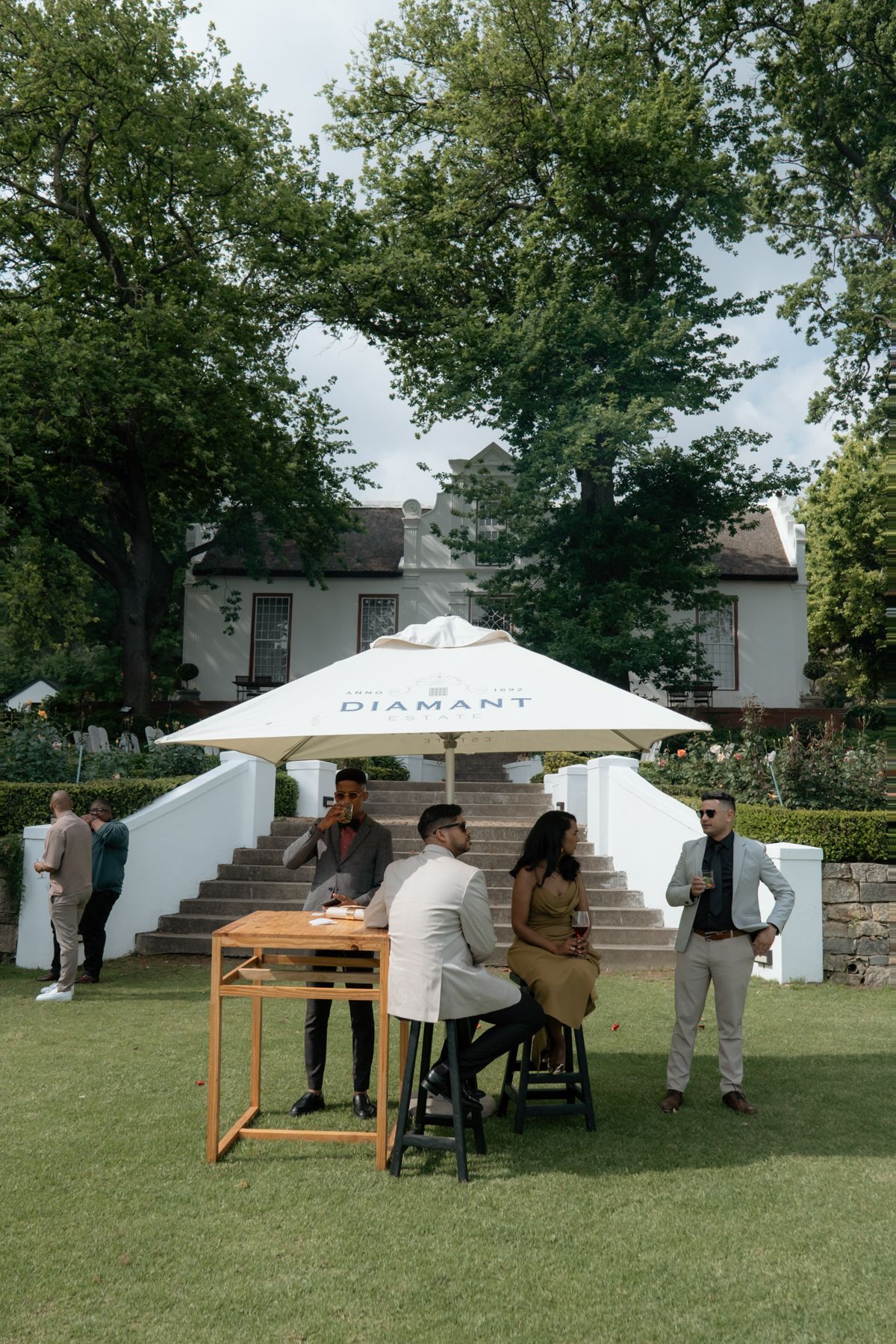 Wedding guests socializing under Diamant branded umbrella on lush green lawn venue.