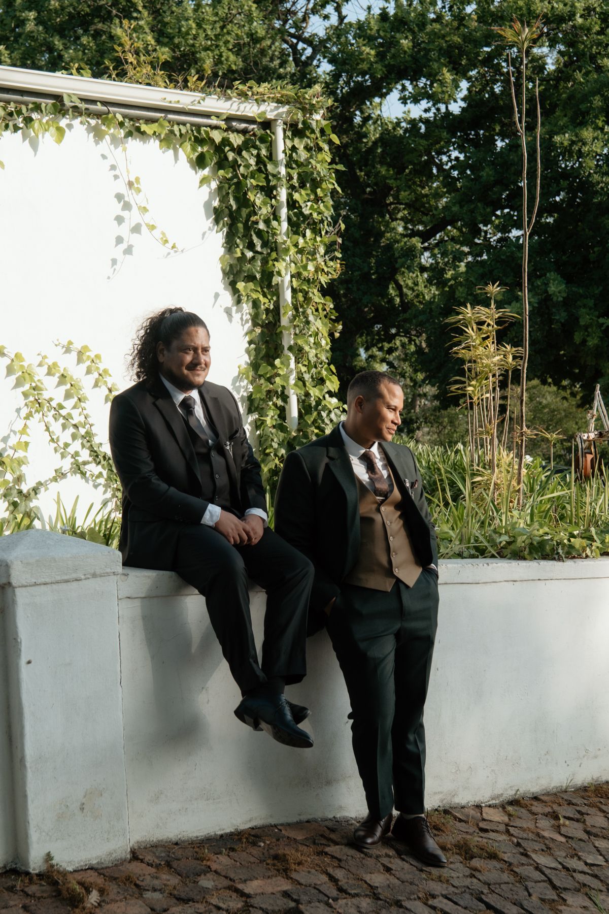 Two groomsmen in dark suits relax and laugh together against white garden wall with ivy backdrop.
