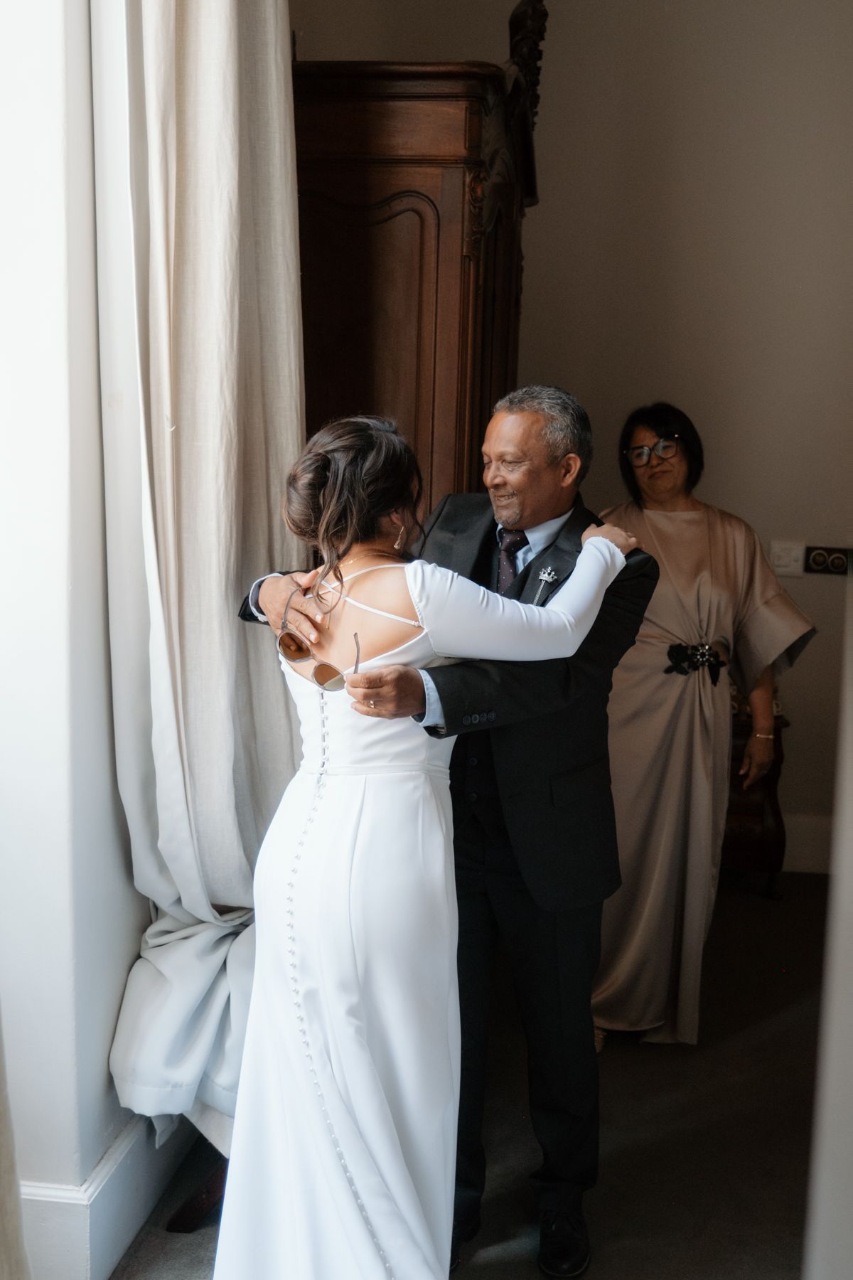 Father embraces bride in elegant backless white wedding gown while mother watches lovingly during bridal preparation.
