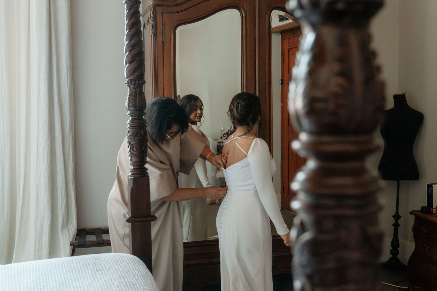Bride in white gown being helped by bridesmaid in front of ornate mirror in elegant bedroom.