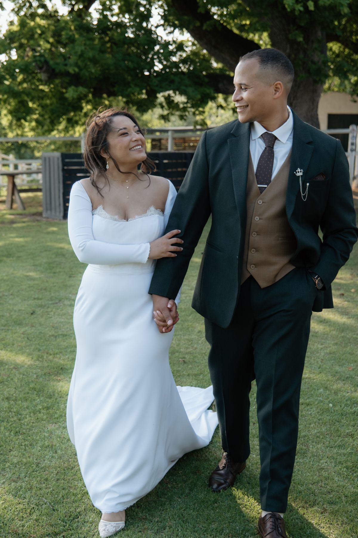 Smiling bride and groom walk hand in hand across green lawn, groom wearing dark suit with brown waistcoat.