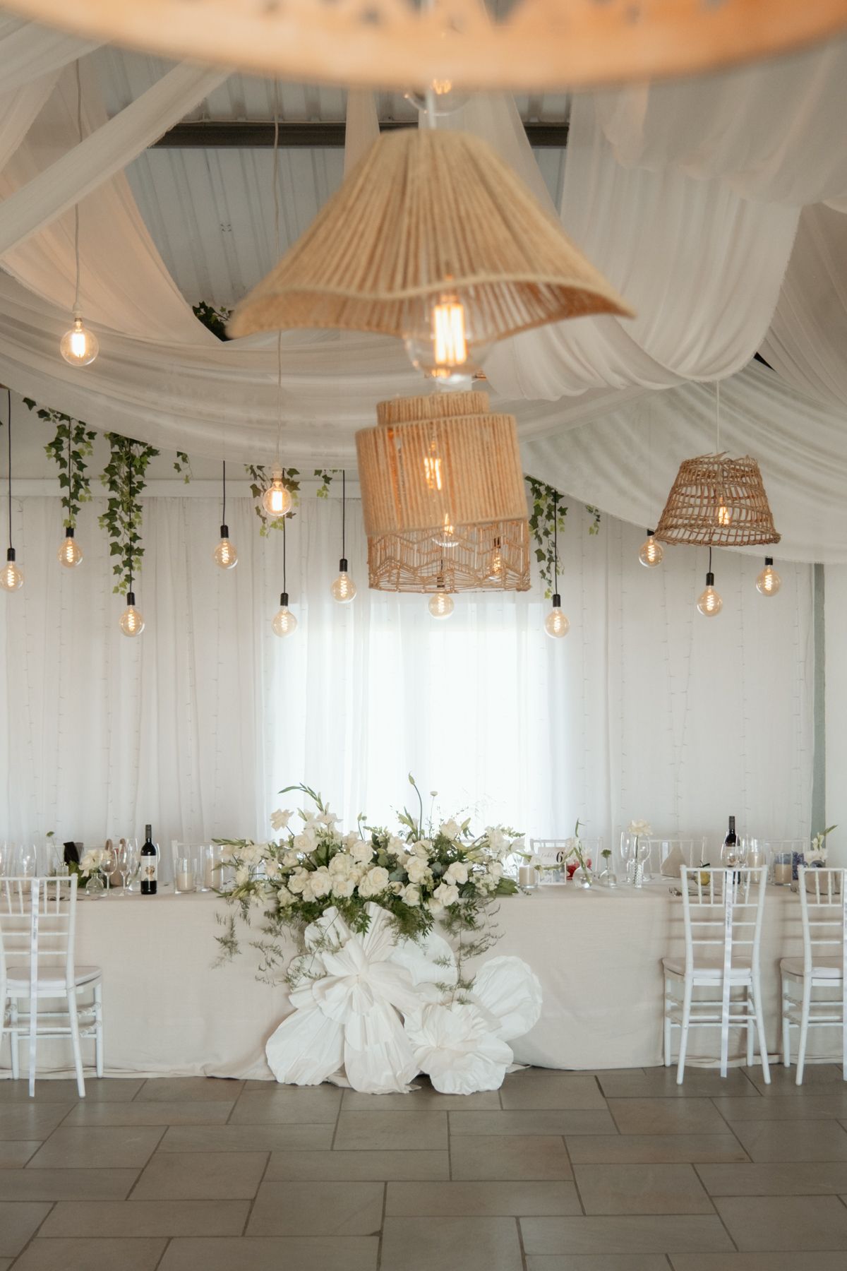 Elegant wedding reception hall with white draping, rattan pendant lights, and floral centerpiece on head table.