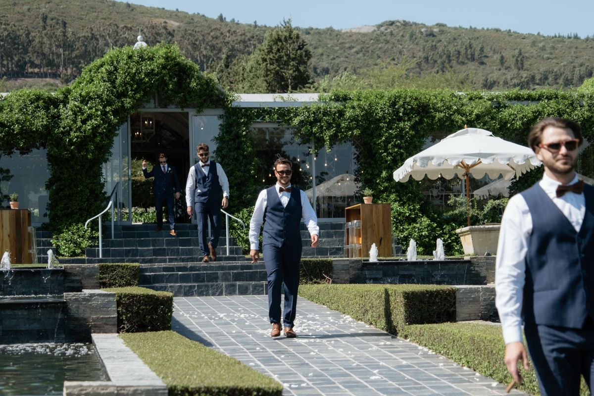 Groomsmen in navy suits walk confidently along a garden pathway at a sunny outdoor wedding venue.