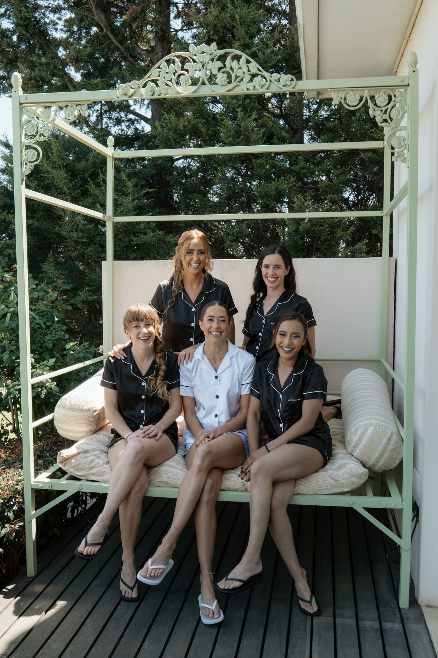 Five women pose together on an ornate white garden daybed, four wearing matching black pajamas and one in white.