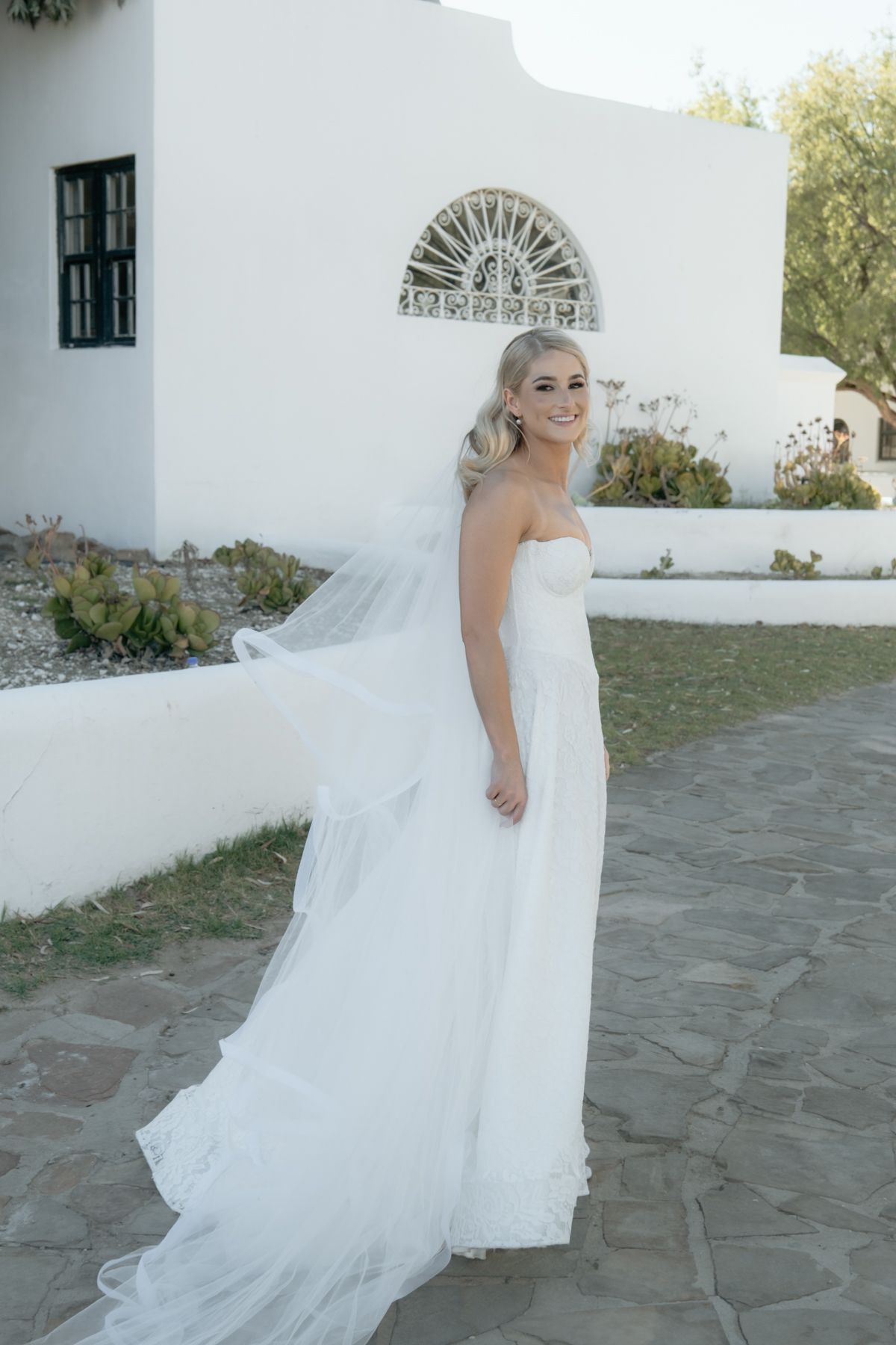 Bride in strapless white gown with flowing veil poses outside white Mediterranean-style building.