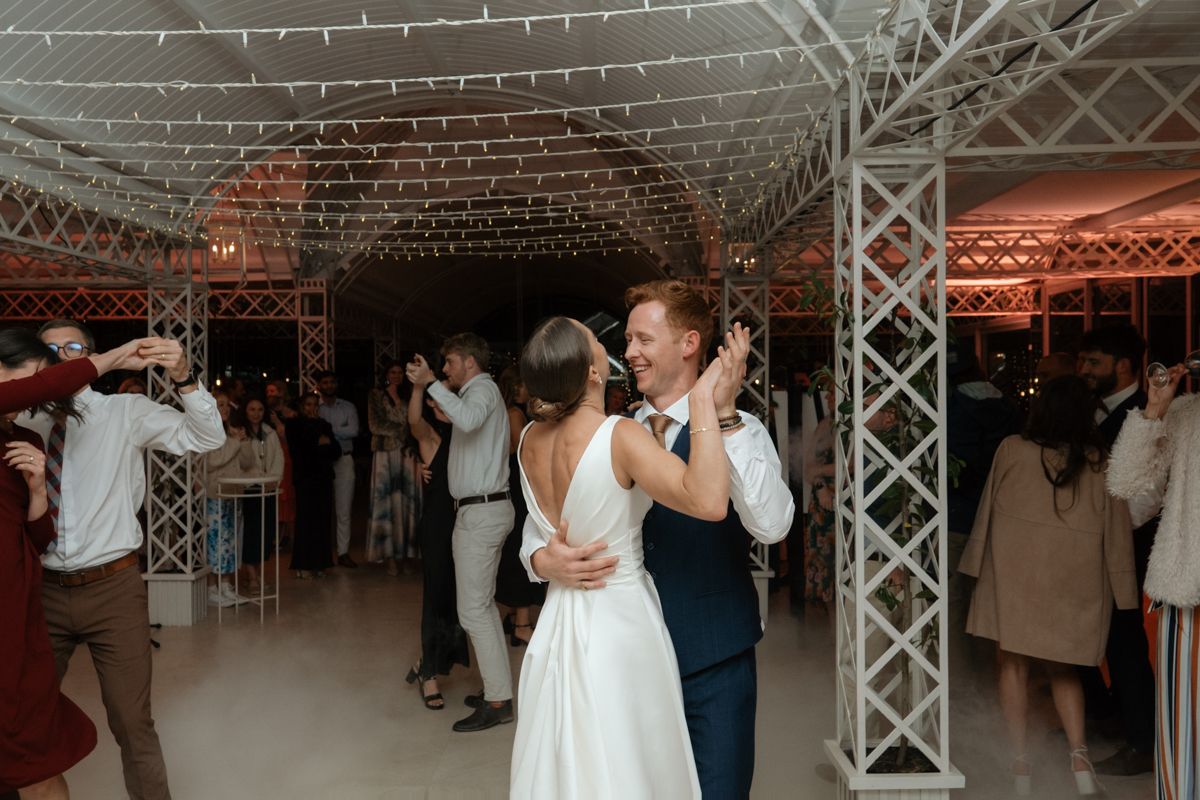 Bride and groom share first dance under twinkling lights at elegant indoor wedding reception with guests dancing.