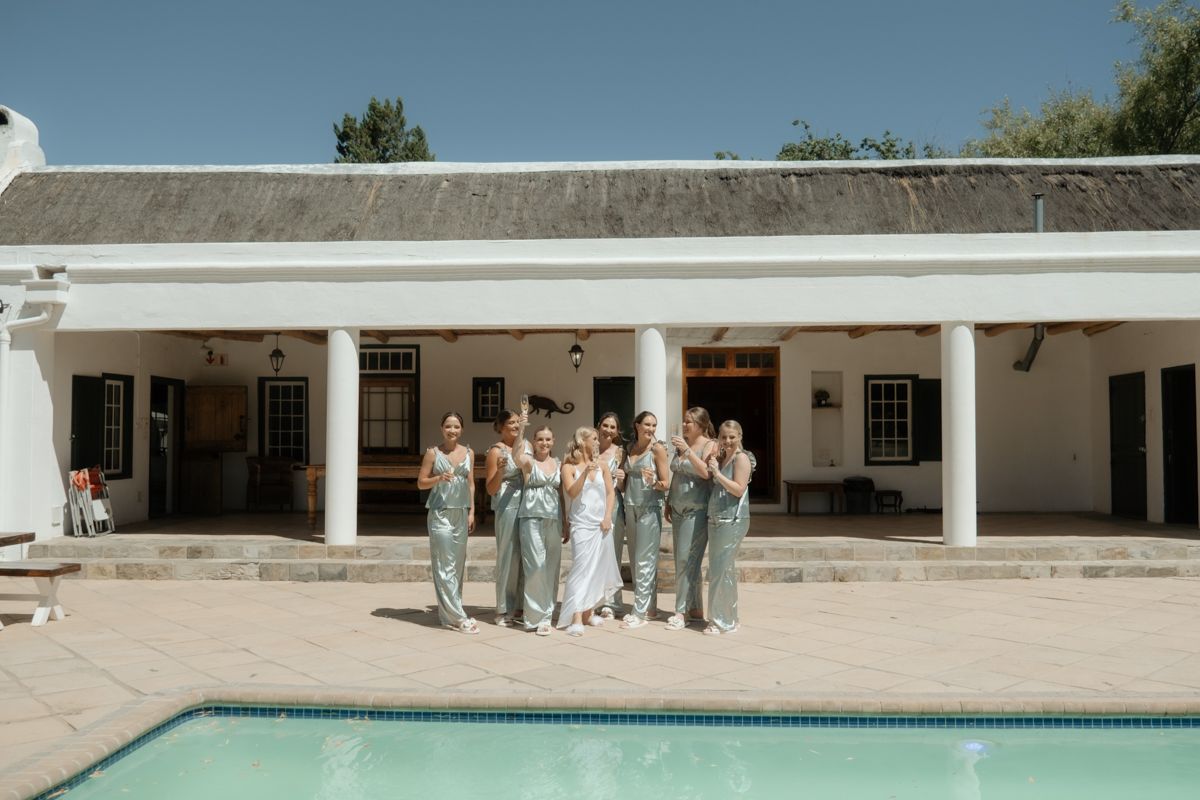 Bride and bridesmaids in sage green dresses posing by a pool at an elegant outdoor wedding venue.