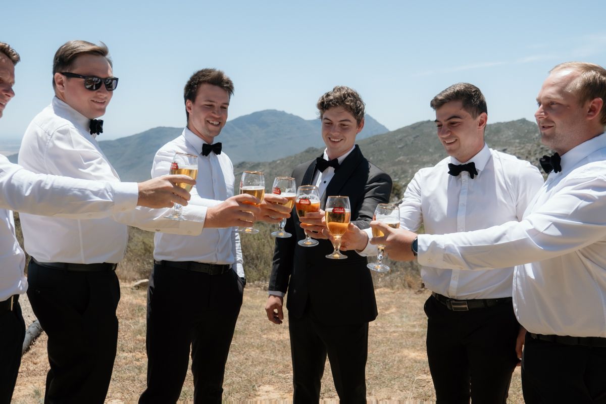 Six groomsmen in black bow ties and white shirts toasting with champagne glasses outdoors with mountains in background.