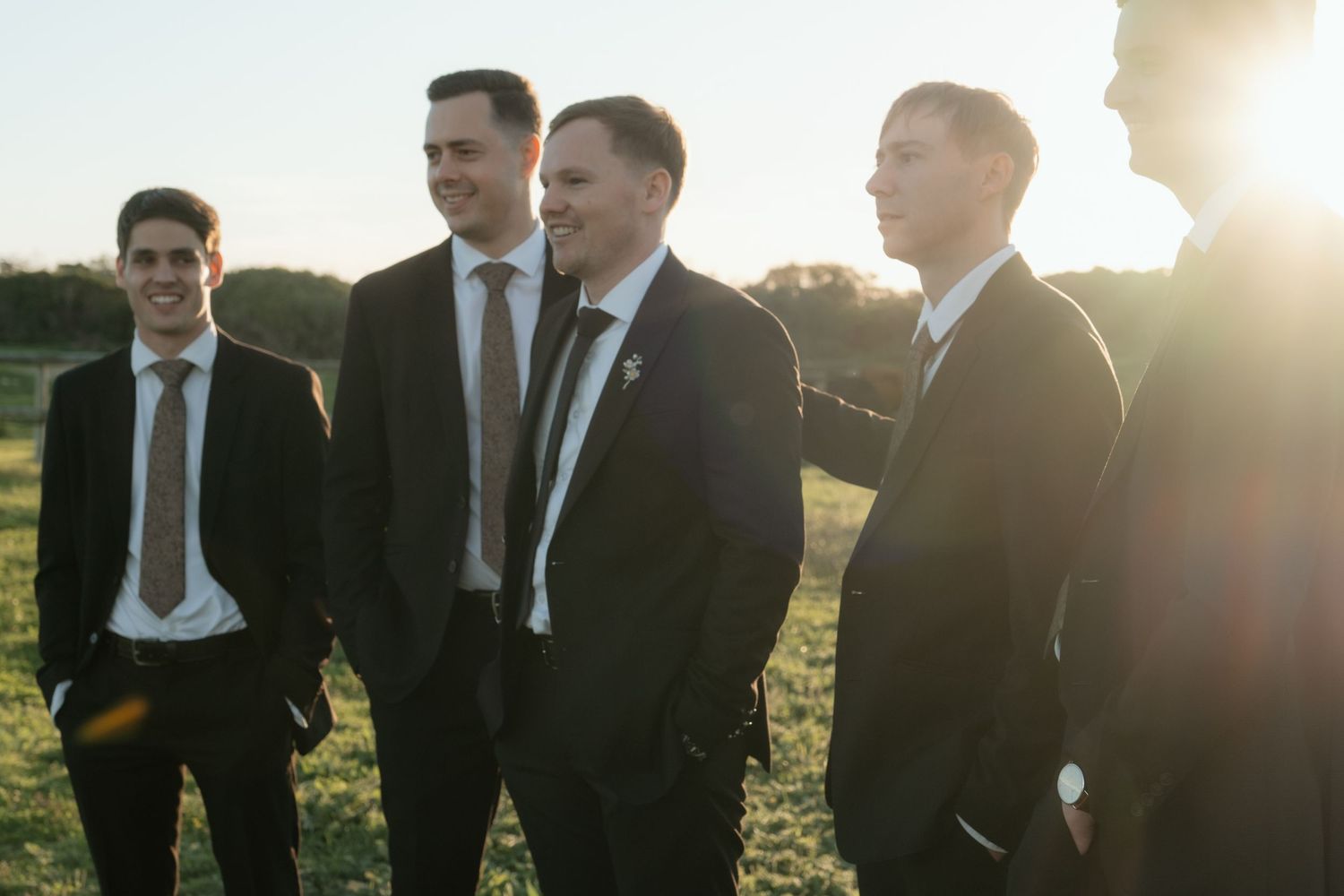 Four groomsmen in dark suits and ties smiling together outdoors in golden sunset light.