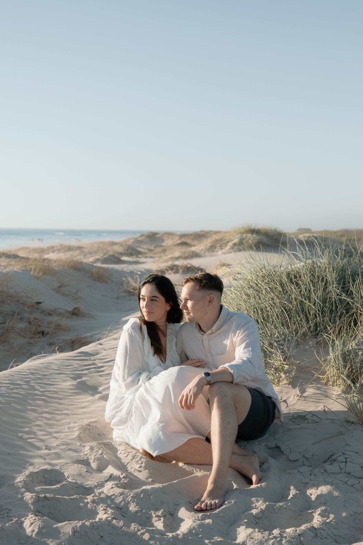 Three romantic photos show a couple sitting together on sandy dunes against a bright sky background.