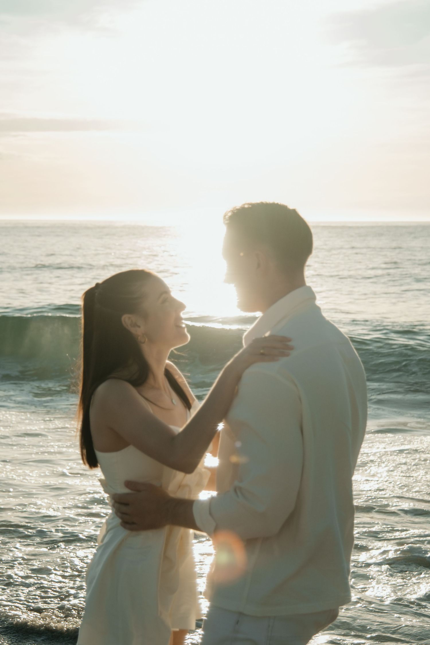 A couple embraces on a beach at sunset with ocean waves and golden sunlight creating a romantic silhouette.