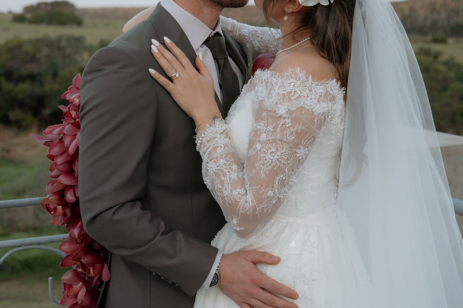 Bride in lace long-sleeve wedding dress embraces groom in gray suit outdoors at romantic wedding ceremony.