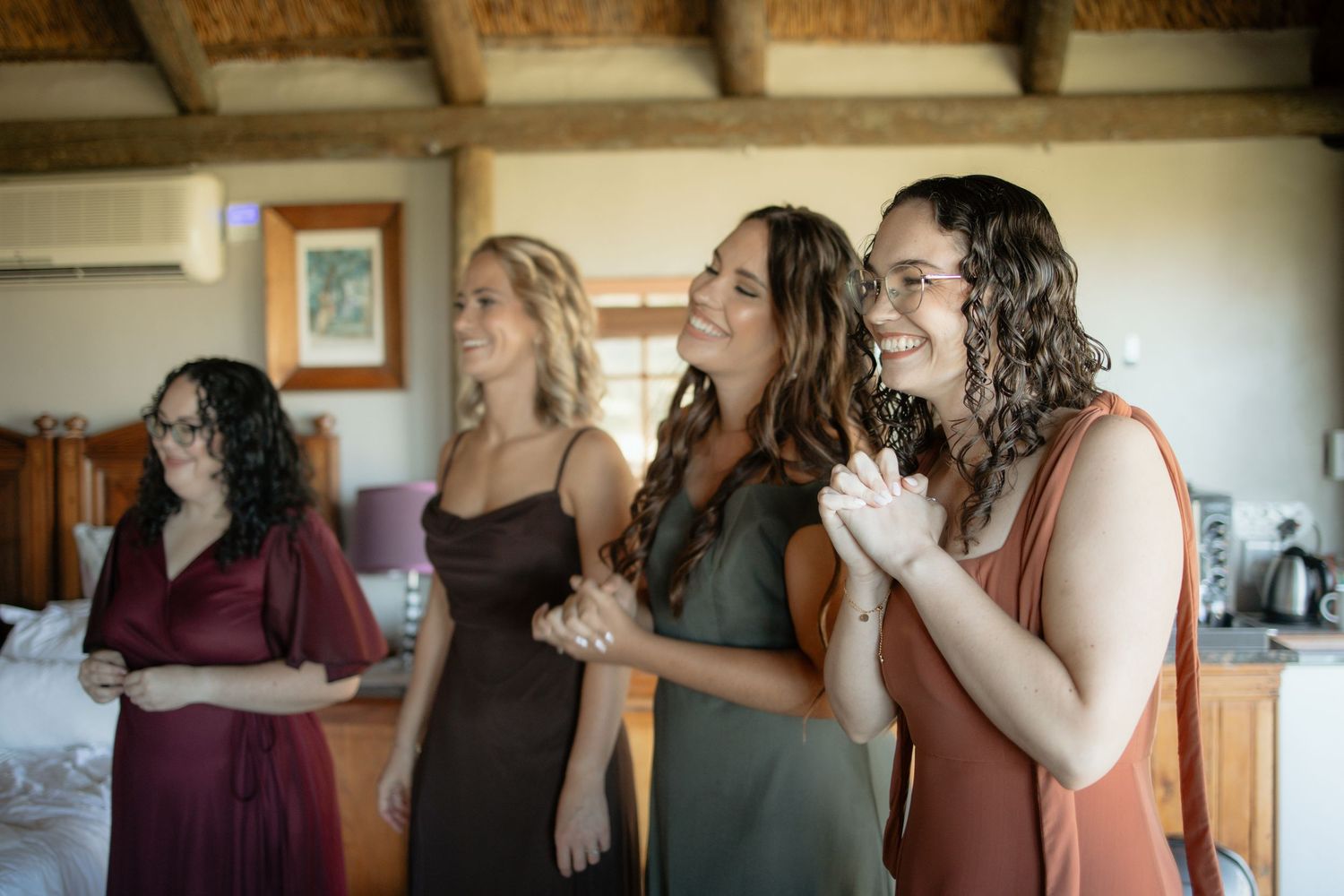 Four bridesmaids in colorful dresses laughing and holding hands joyfully during wedding preparations indoors.