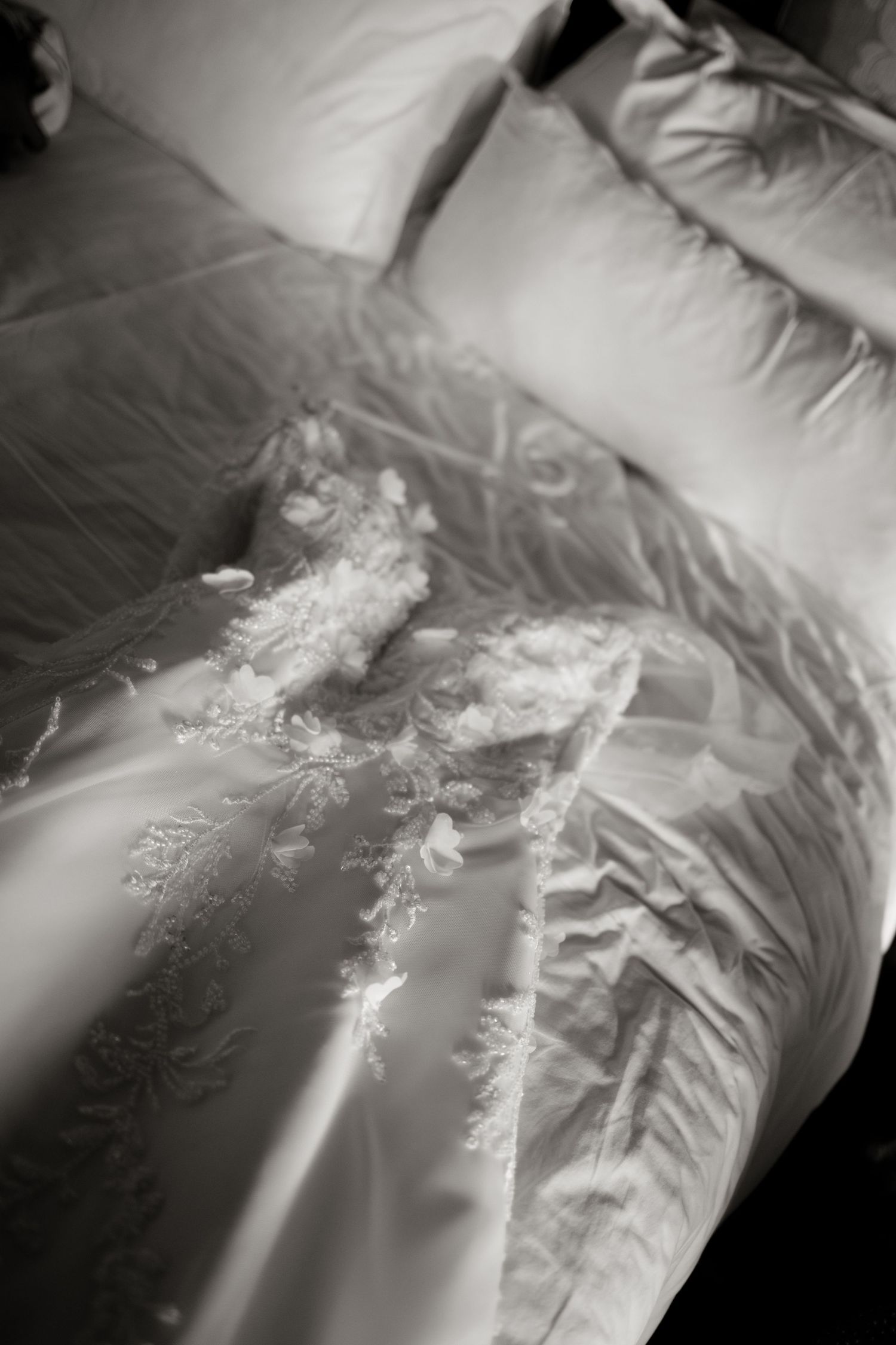 Close-up black and white detail of lace embroidered wedding dress laid on satin bed.