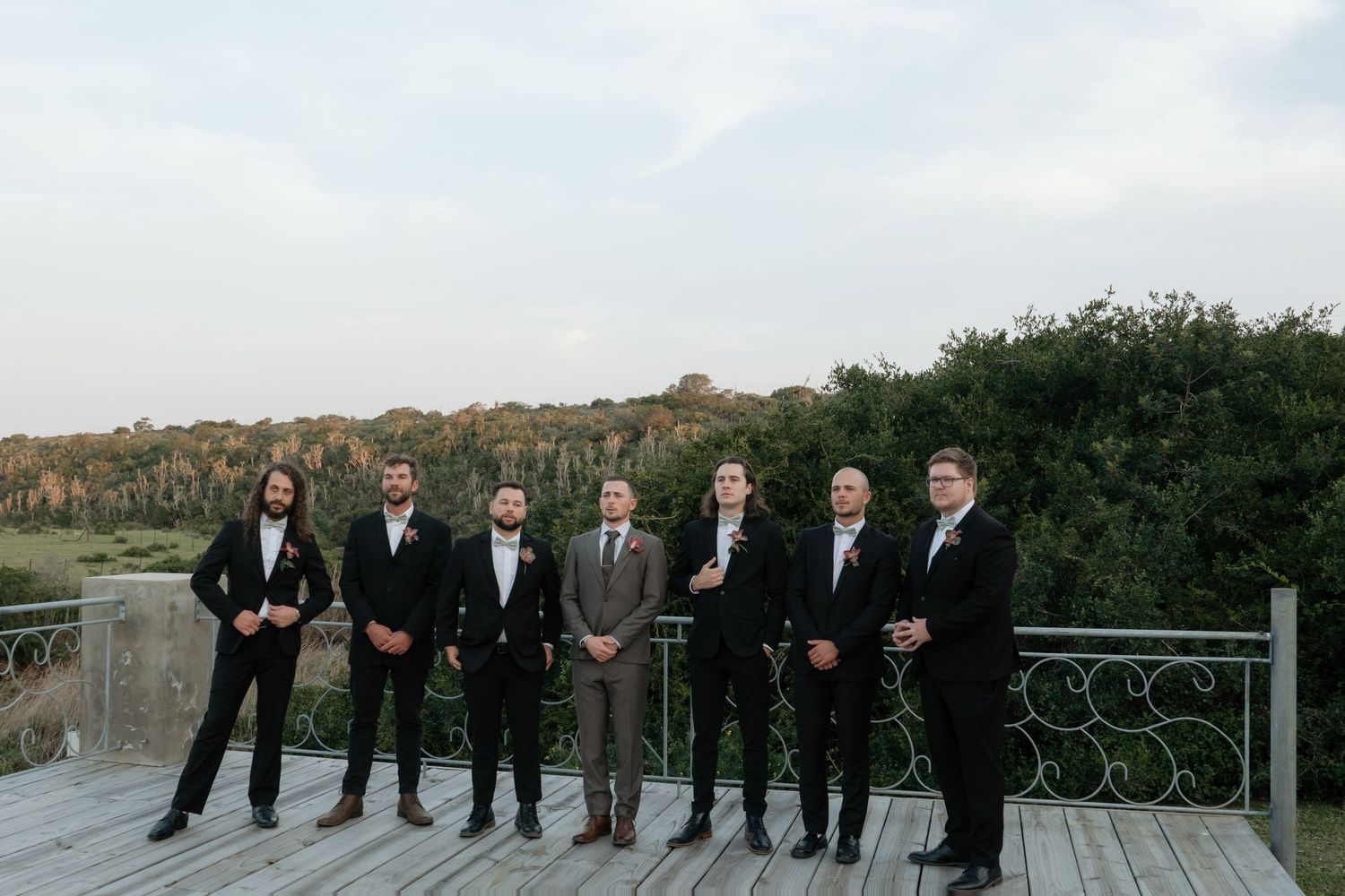 Seven groomsmen in black tuxedos and one in brown suit pose on wooden deck with lush green trees backdrop.