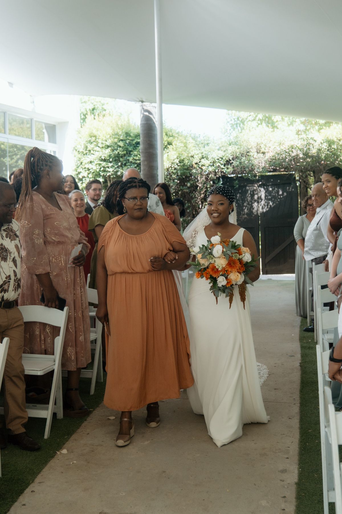 Bride in white gown walks down aisle with escort at outdoor garden wedding ceremony.