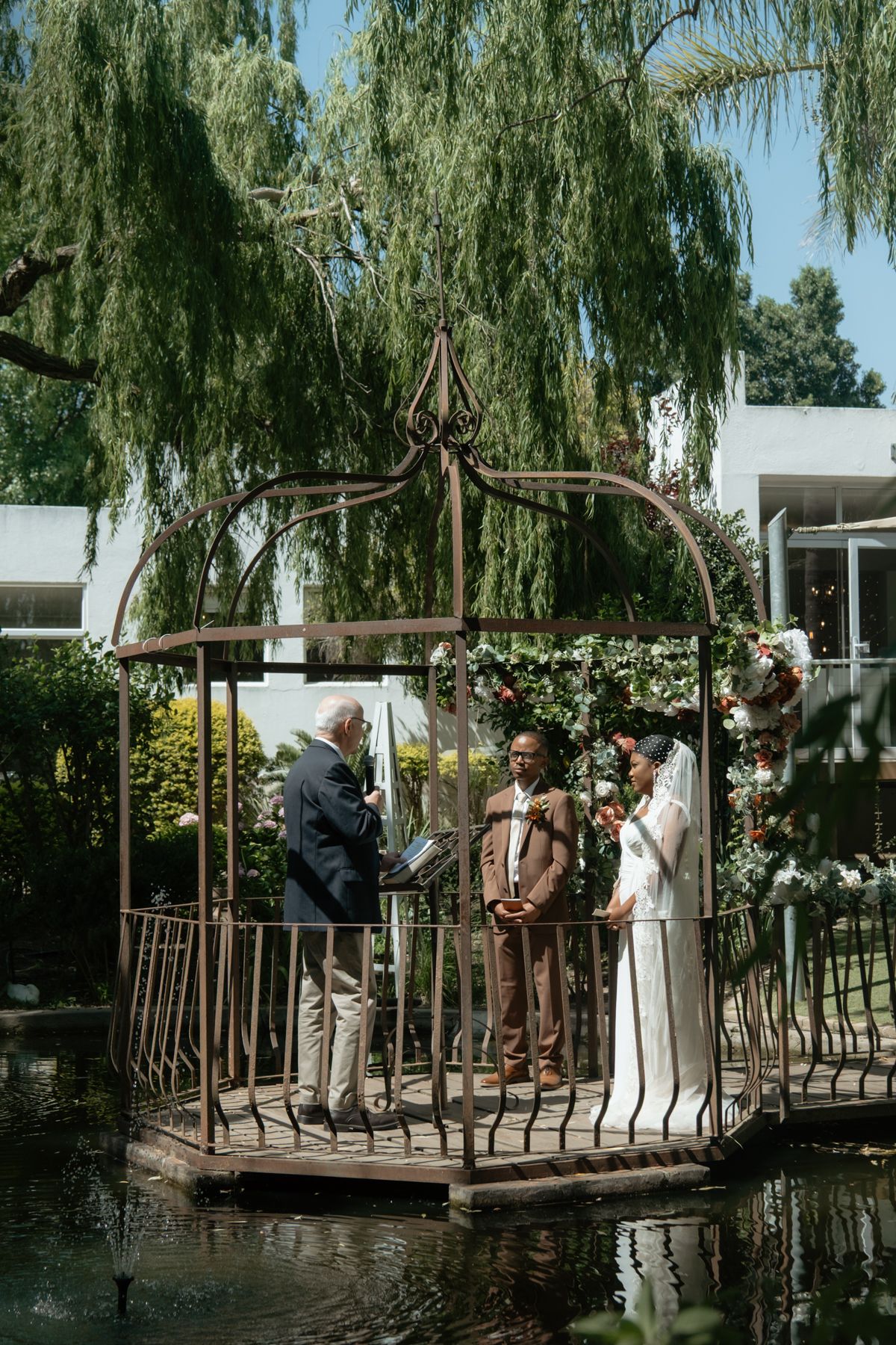Couple exchanges vows with officiant in rustic iron gazebo over pond at lush outdoor garden wedding.