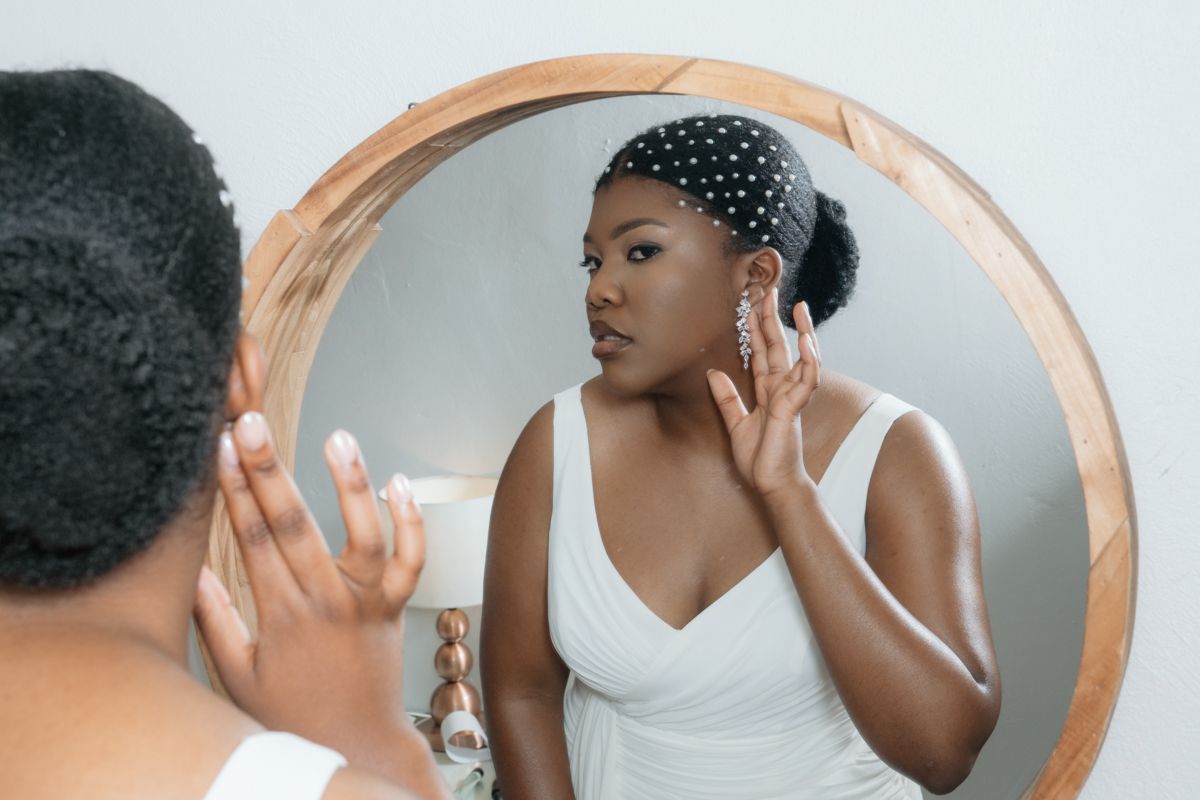 Black woman in white dress applying skincare while looking at her reflection in a round wooden mirror.