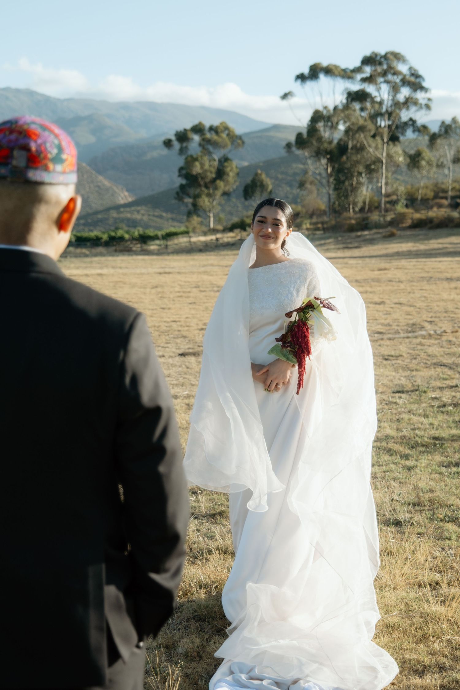 Bride in white gown with veil holds bouquet facing groom in scenic mountain landscape.