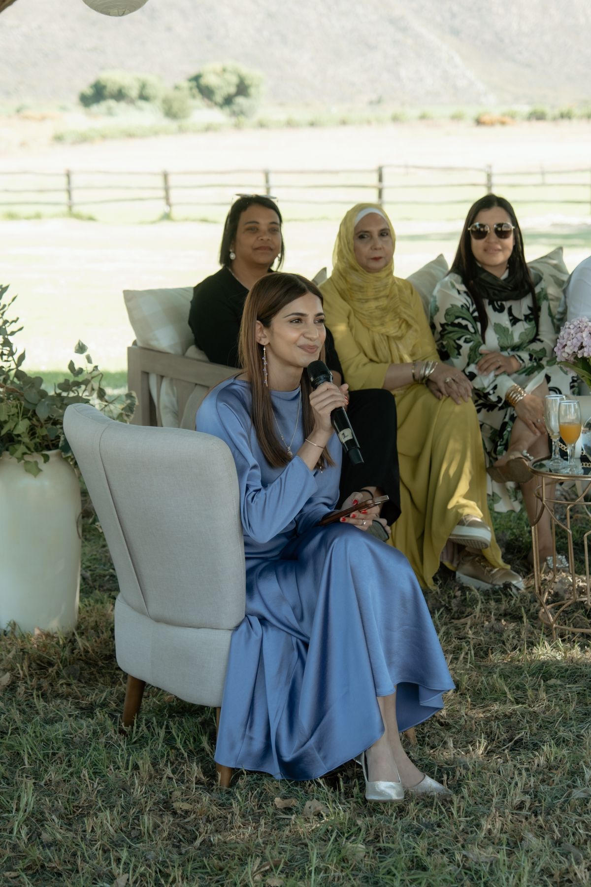 Woman in blue dress holds microphone while seated at outdoor wedding with guests behind her.