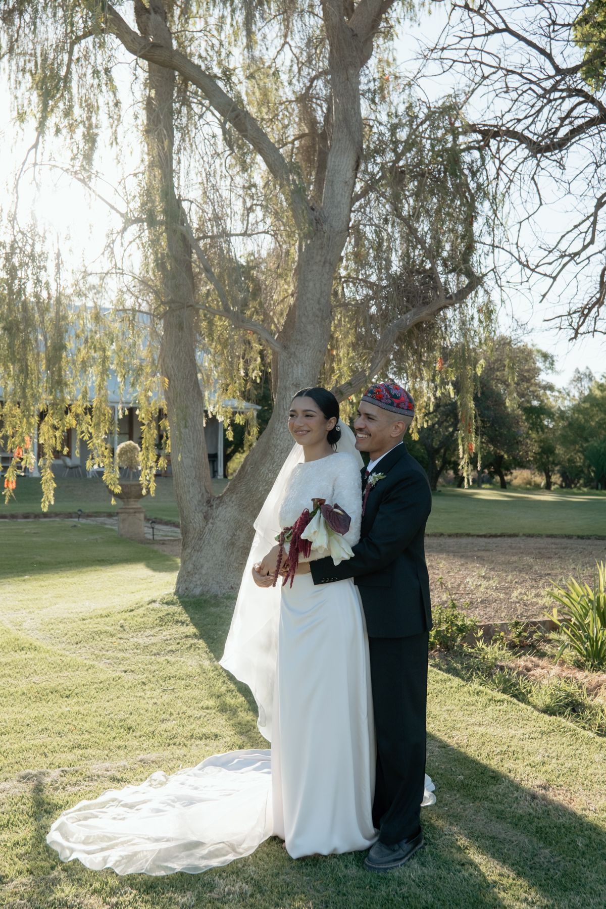Groom in black suit and kippah embraces bride in white gown under a willow tree outdoors.