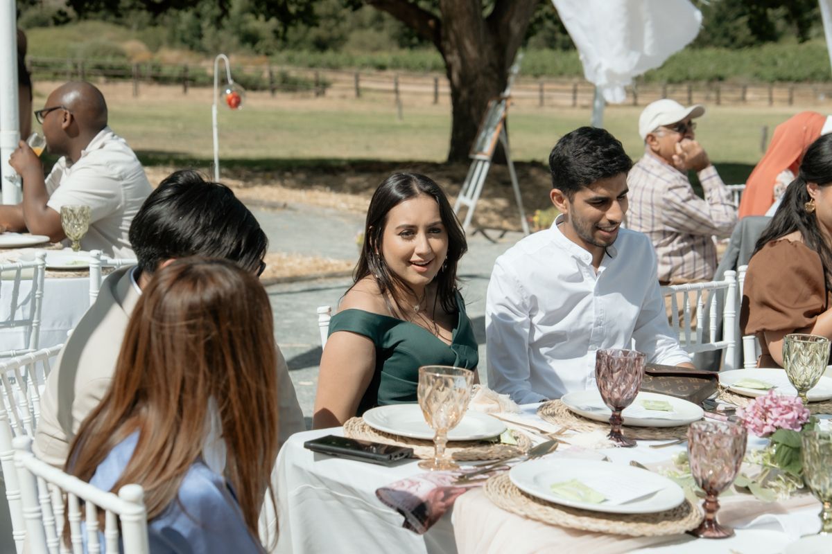Guests in formal attire enjoy conversation at outdoor wedding reception table with pink glassware.