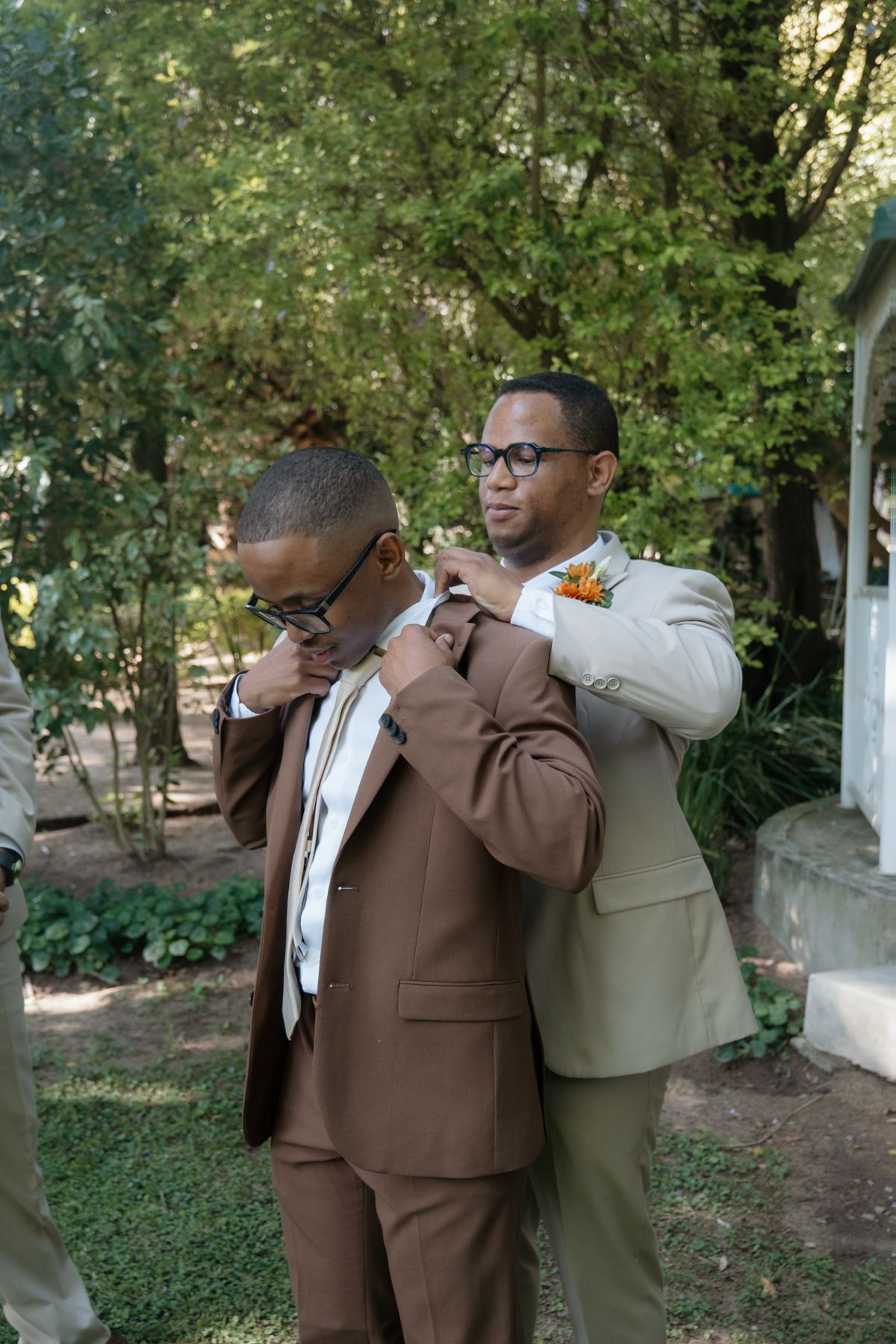 A groomsman in a beige suit adjusts the collar of a groom in a brown suit outdoors.